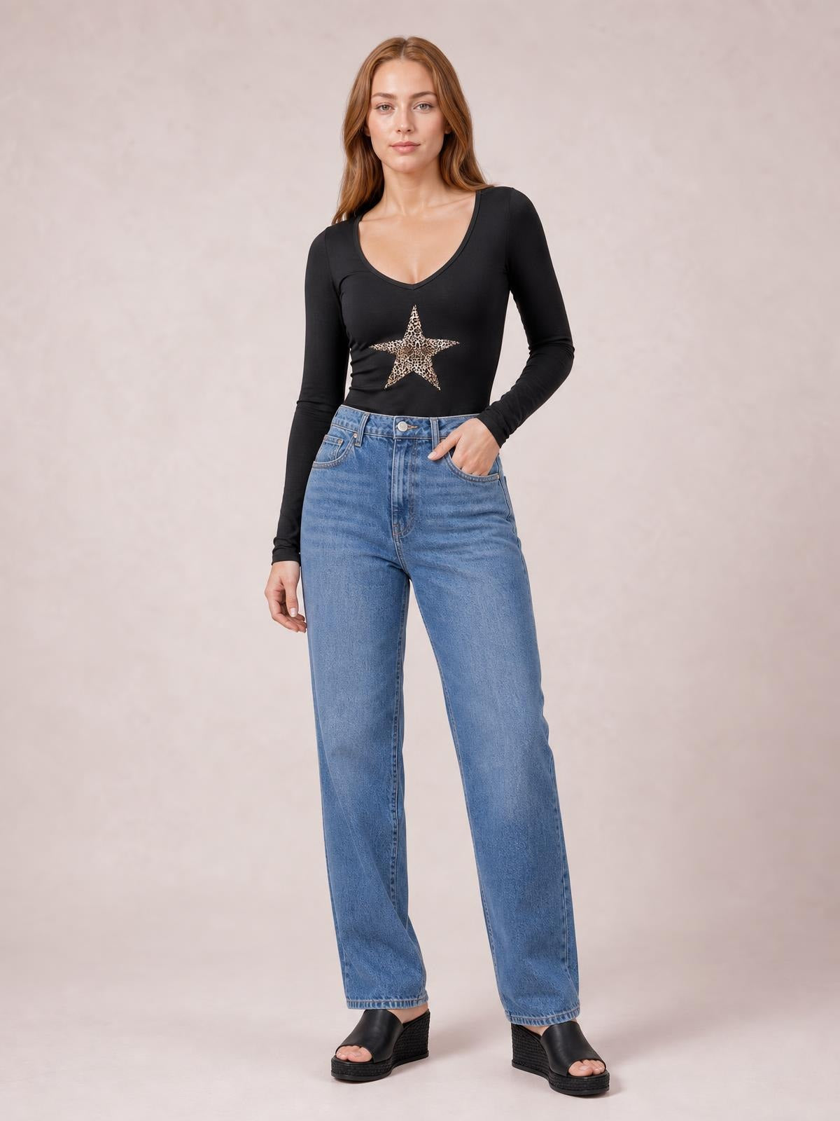 Model stands in a black, fitted long sleeve T-shirt with a V-neck and leopard-print star graphic, one hand resting in her jeans pocket.