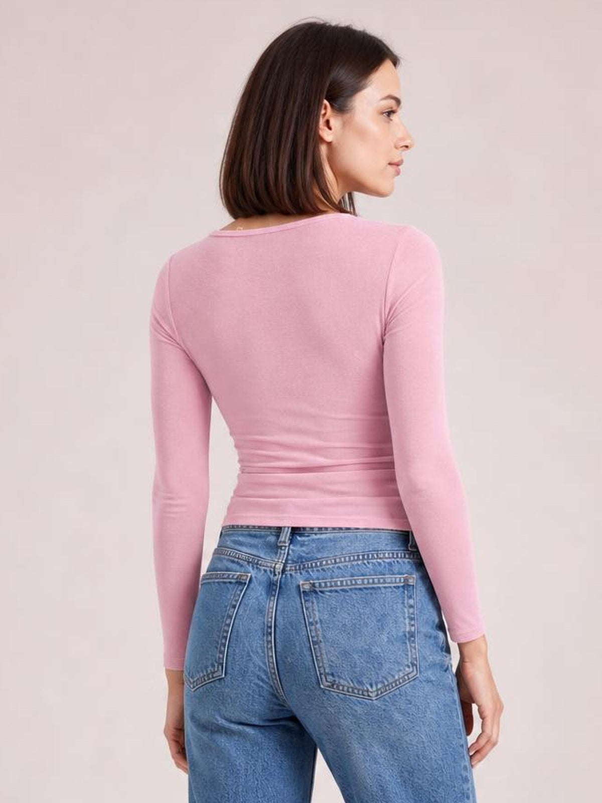 A model turns to show the back of a fitted pink long sleeve T-shirt with a round neckline, smooth stretch construction, and hip-length silhouette.