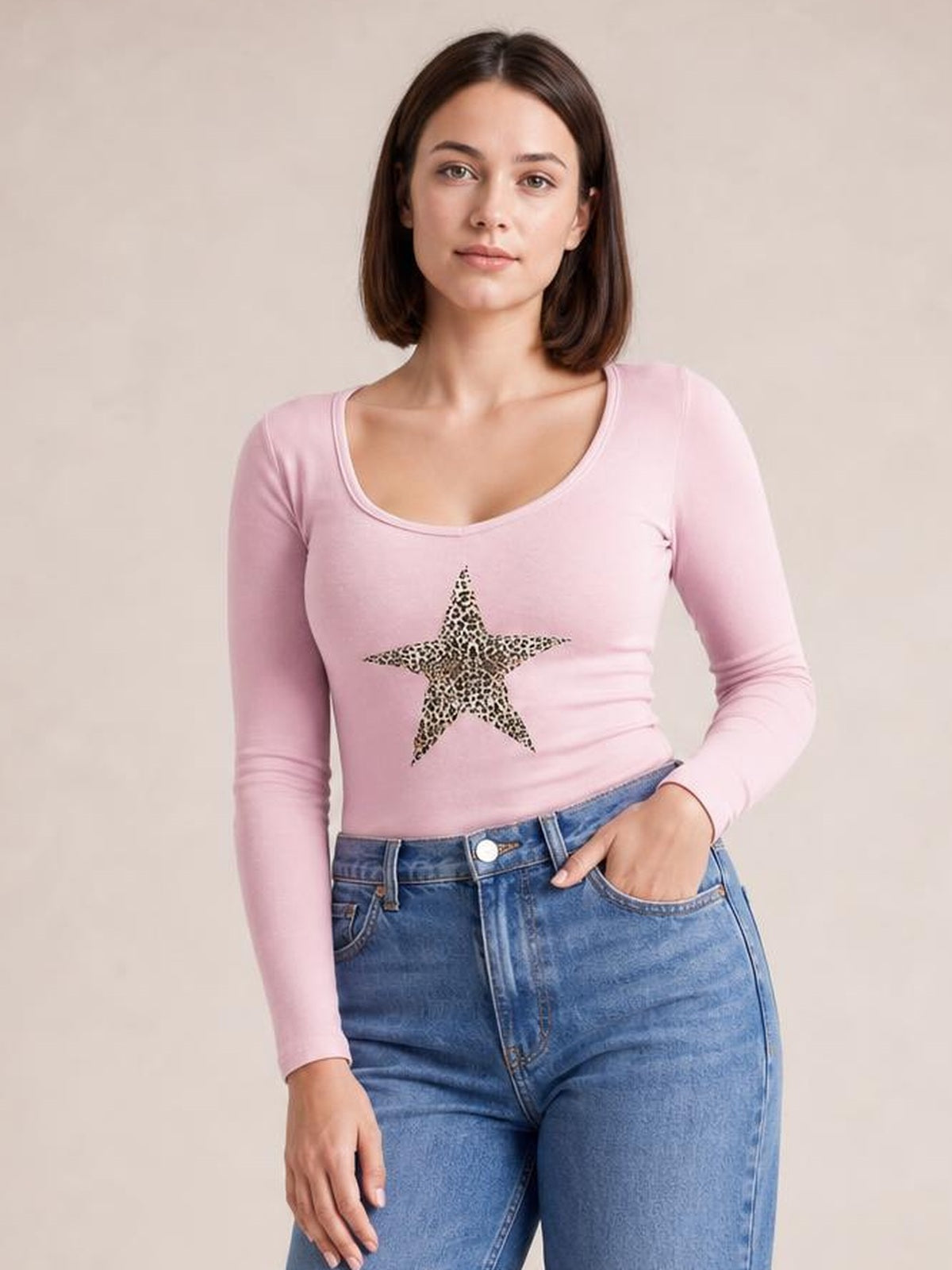 A model wears a fitted pink long sleeve T-shirt with a scoop neckline and bold leopard-print star graphic, posing with one hand in her jeans pocket.
