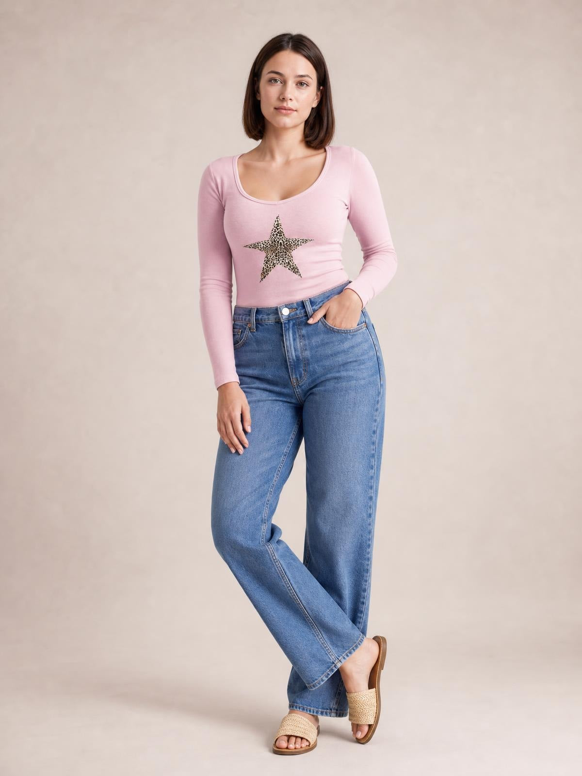 A model wears a pink Leopard Star long sleeve T-shirt with a scoop neckline, fitted stretchy silhouette, and leopard-print star graphic, posing with crossed legs.