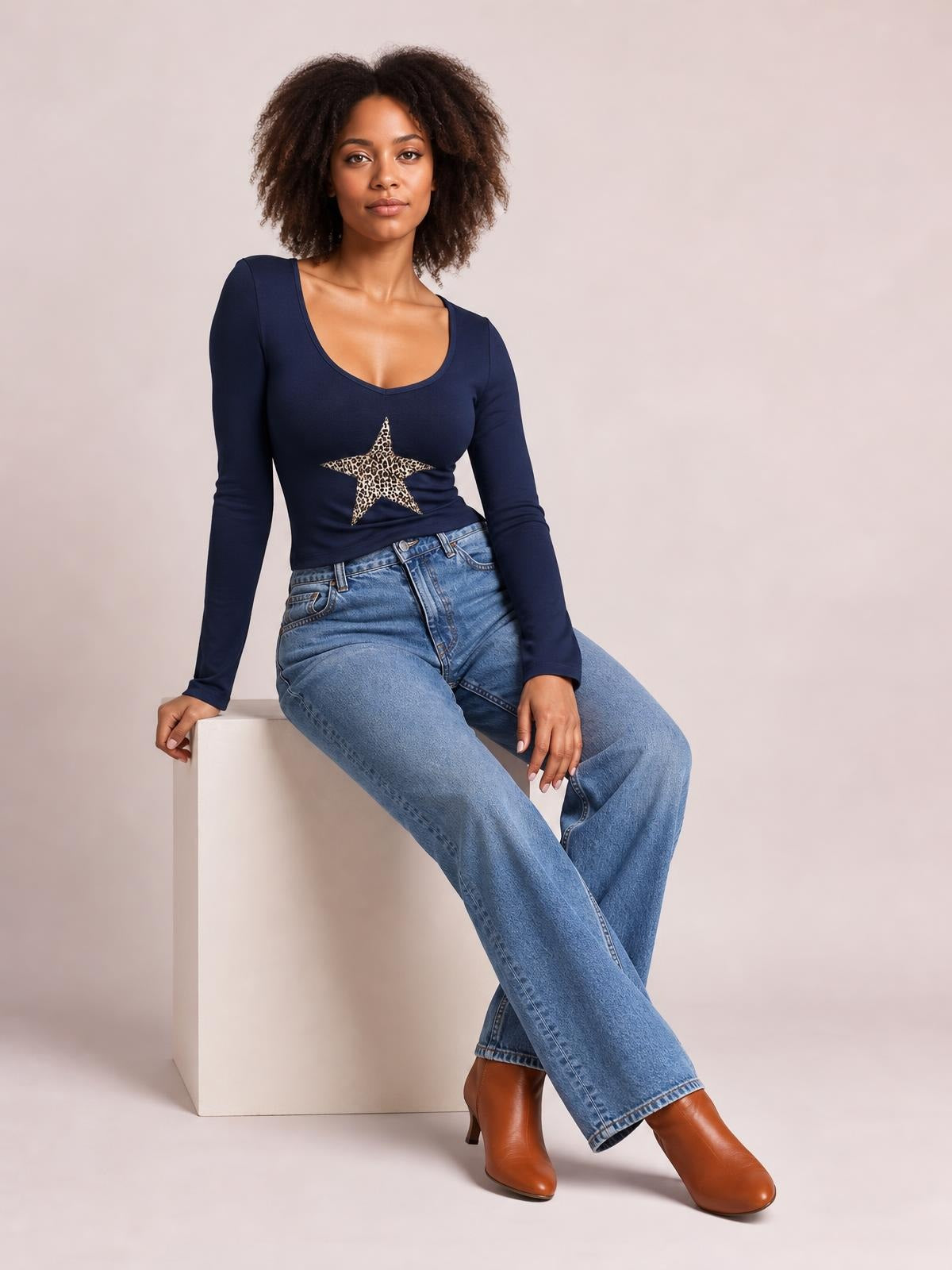 A model sits with crossed legs wearing a navy long sleeve T-shirt with a scoop neckline, fitted silhouette, and bold leopard-print star graphic.