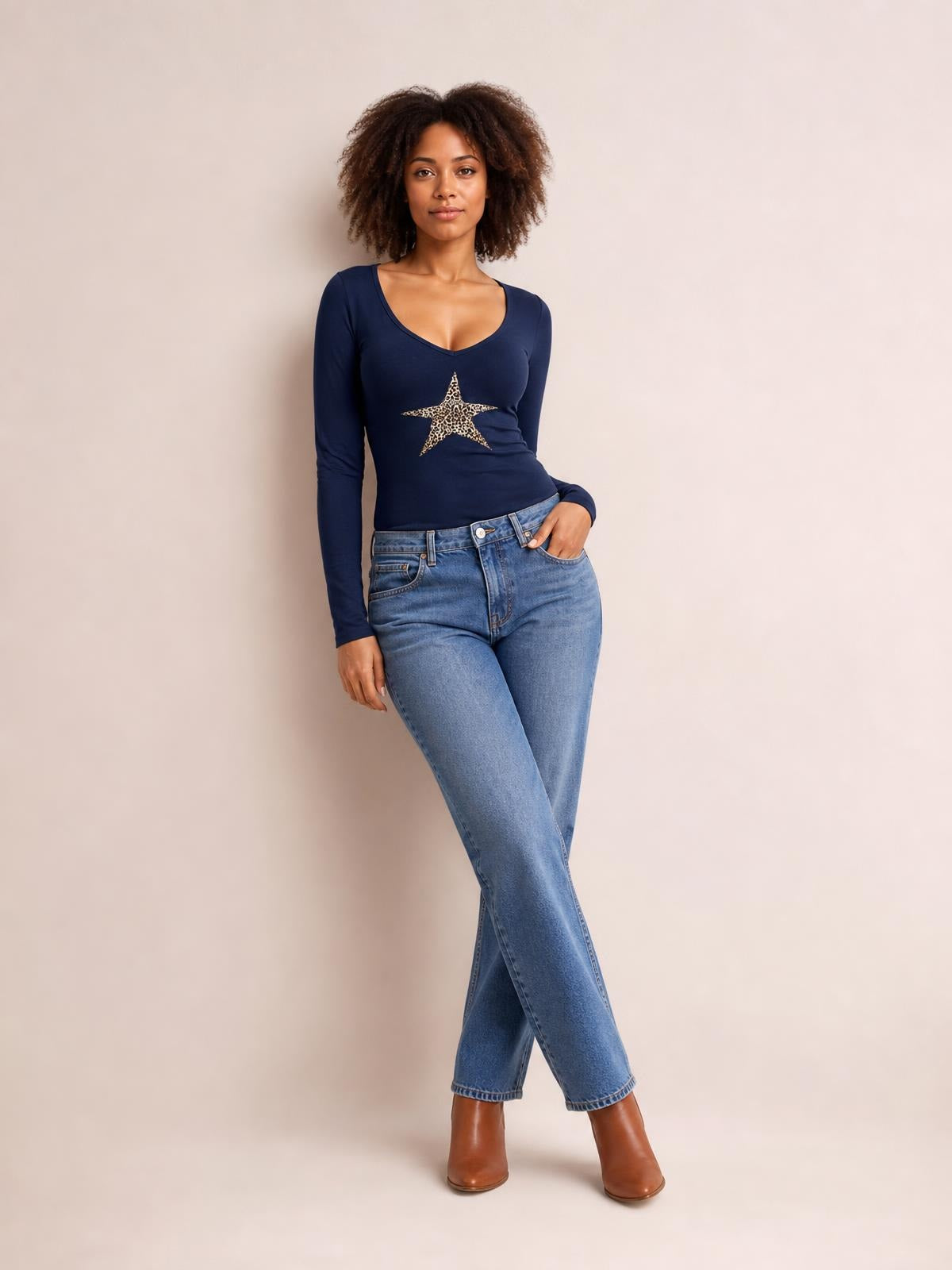 A model poses in a fitted navy Leopard Star long sleeve T-shirt with a scoop neckline and leopard-print star graphic, one hand in her jeans pocket and legs crossed.