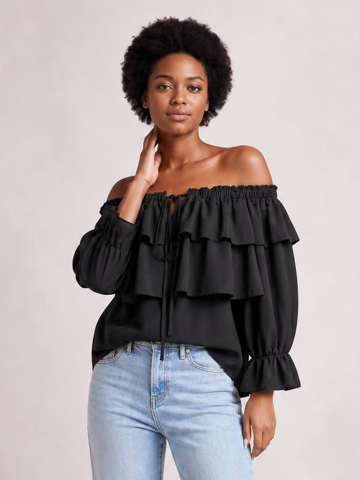 Model poses with one hand at her neck wearing a black off-shoulder blouse with layered ruffles, voluminous flounce sleeves, elastic neckline, and drawstring detail.