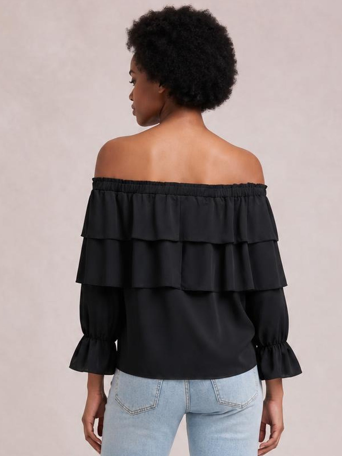 Model shown from the back wearing a black off-shoulder blouse with layered ruffles, flounce sleeves, gathered cuffs, and a relaxed silhouette.
