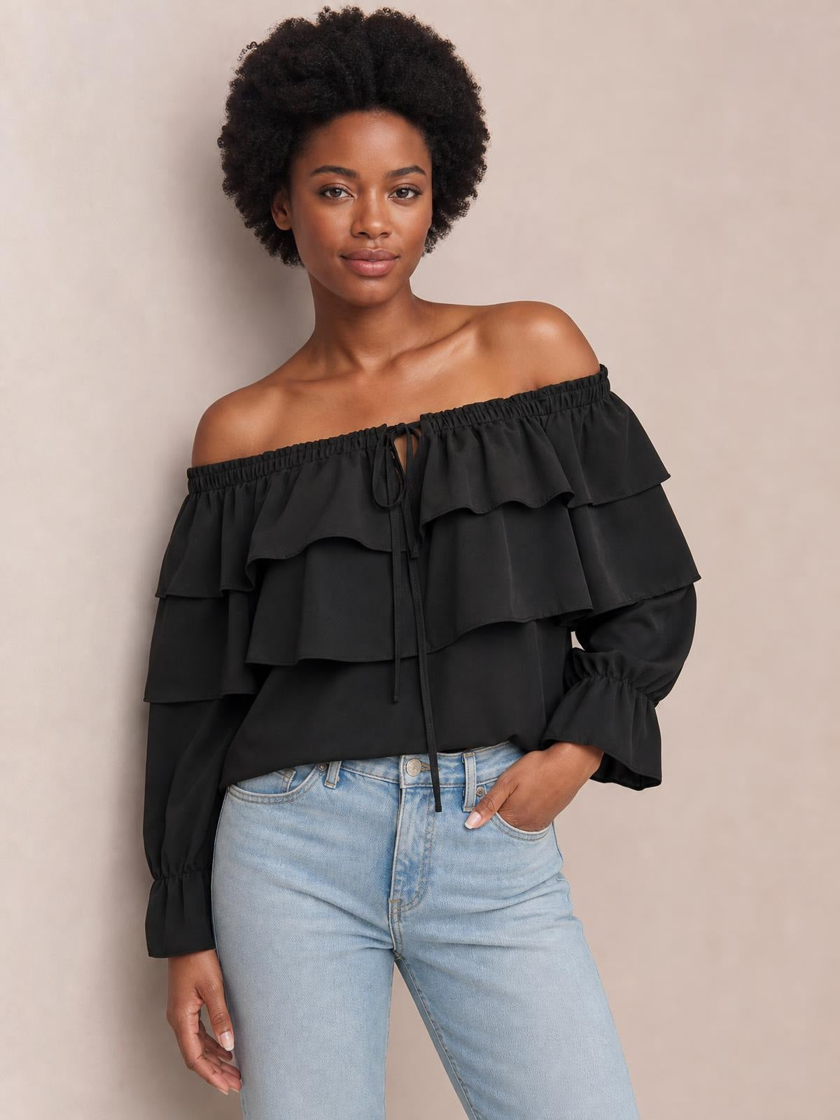 Model poses in a black off-shoulder blouse with layered ruffled flounce sleeves, an elastic neckline, and a front drawstring tie.