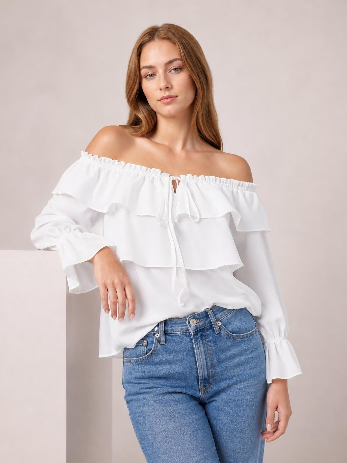 Model wears a white off-shoulder blouse with layered ruffled flounce sleeves, a drawstring neckline, and a relaxed silhouette, posing with one hand resting on a ledge.