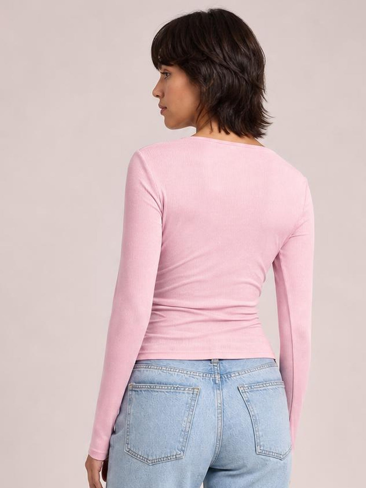 Model shown from the back in a fitted pink long-sleeve T-shirt with a smooth ribbed texture and gently ruched silhouette, turning in profile.