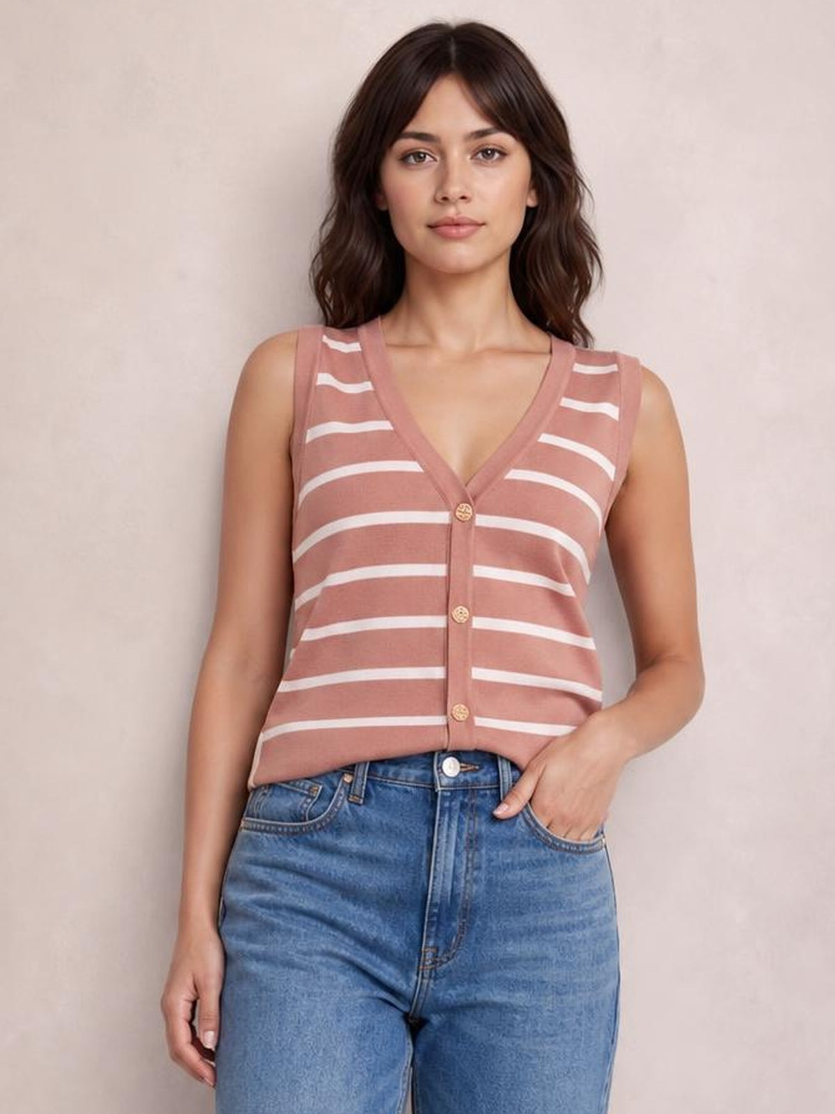 Model wearing a dusty rose striped V-neck knit tank with a button-front design and fitted sleeveless silhouette, posing with one hand in her jeans pocket.