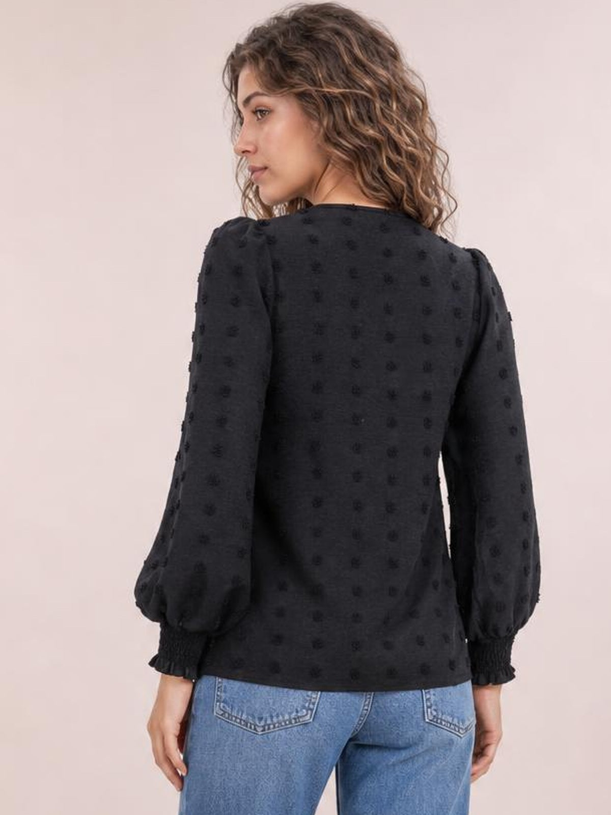 A model looks over her shoulder, showing a black Swiss dot blouse with a relaxed silhouette, long puff sleeves, and ruffled cuffs.