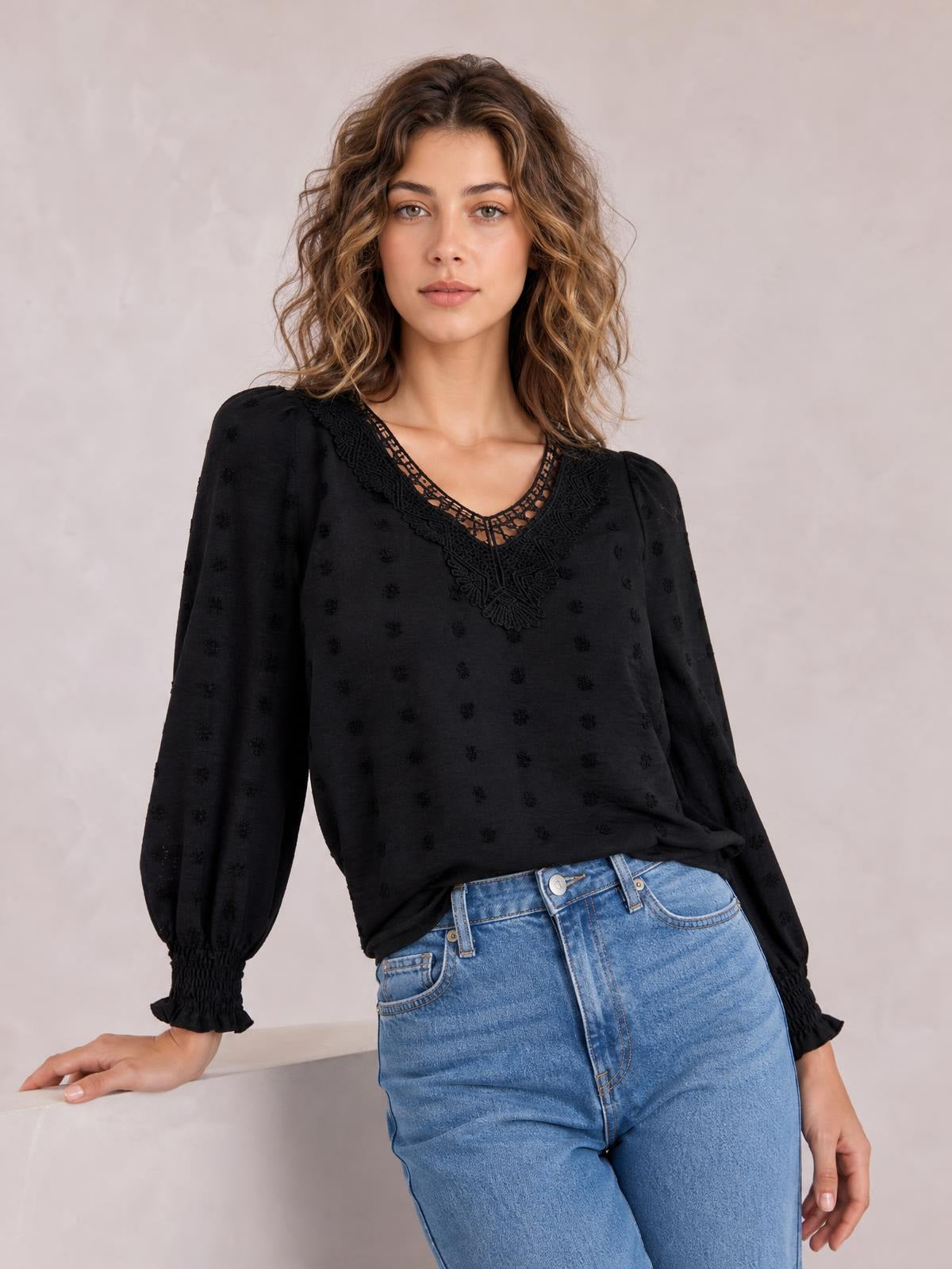 Model wearing a black Swiss dot V-neck blouse with lace neckline detail, long sleeves, gathered cuffs, and a relaxed silhouette while leaning casually.