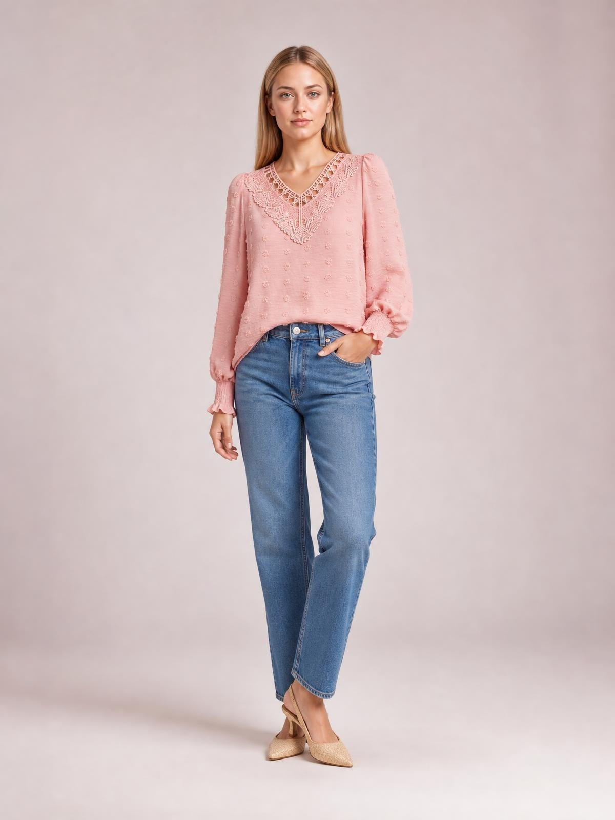 A model wears a pink Swiss dot blouse with a lace-detail V-neck, long sleeves, gathered cuffs, and a relaxed silhouette, posing with one hand in her jeans pocket.