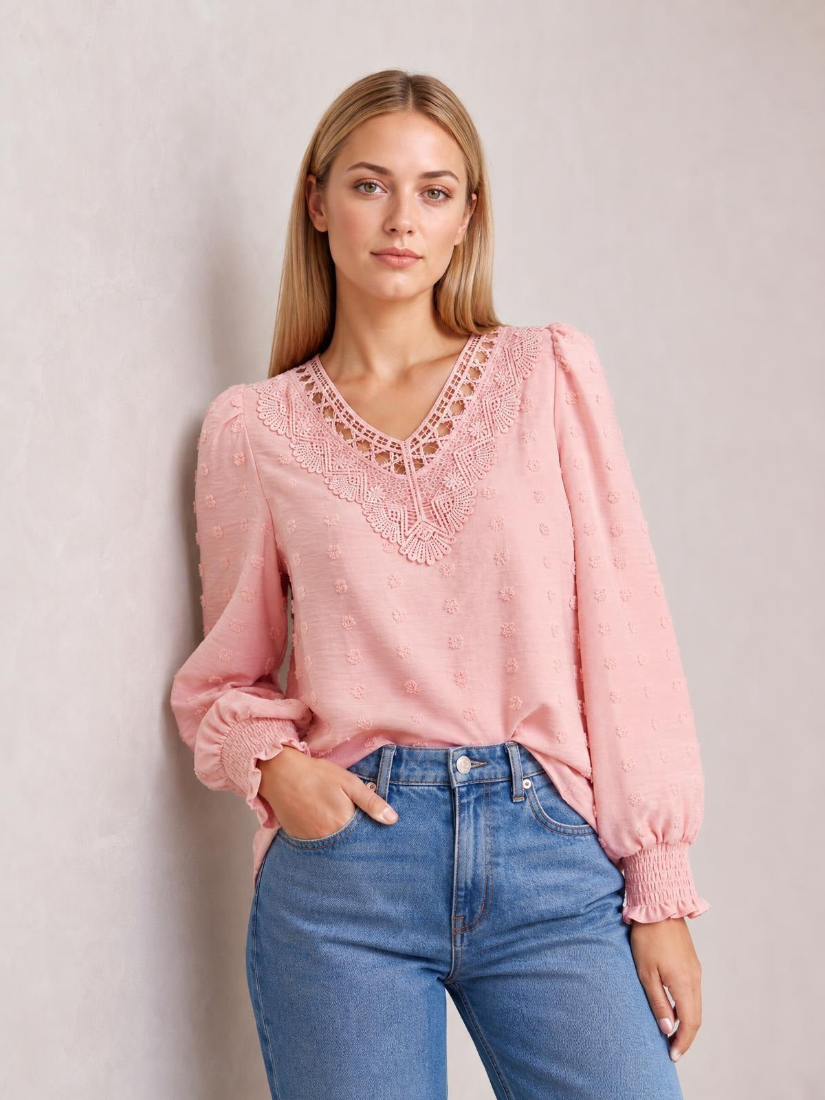 A model poses with one hand in her jeans pocket, wearing a pink Swiss dot blouse with a V-neck, ornate lace detail, long sleeves, and gathered cuffs.