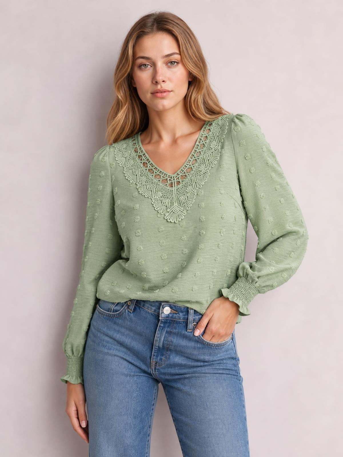 A model poses in a sage green Swiss dot blouse with a V-neck, ornate lace detail, long sleeves, gathered cuffs, and a relaxed silhouette, one hand in her jeans pocket.