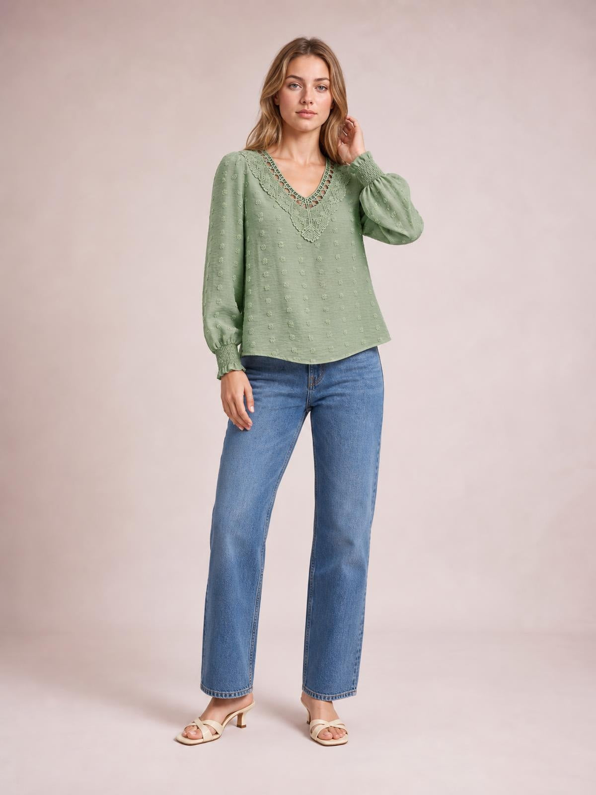 Model wearing a sage green Swiss dot blouse with a lace-trimmed V-neck, semi-sheer fabric, long gathered sleeves, and a relaxed silhouette, posing with one hand in her hair.