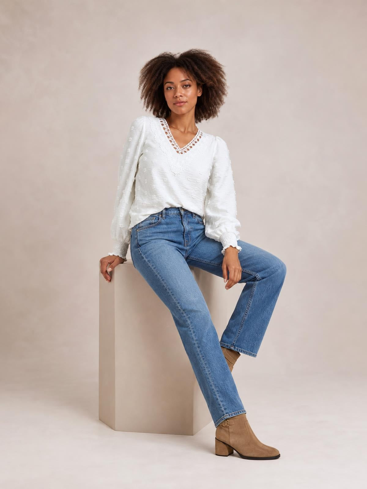 A seated model wears a white Swiss dot blouse with a V-neck lace-trim detail, long sleeves, gathered cuffs, and a relaxed silhouette.