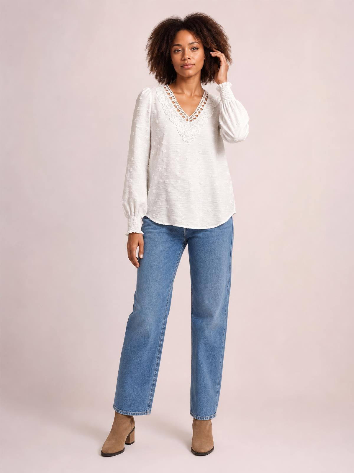 A model stands in a white Swiss dot blouse with a lace-detail V-neck, long sleeves, relaxed silhouette, and curved hem, touching her hair.