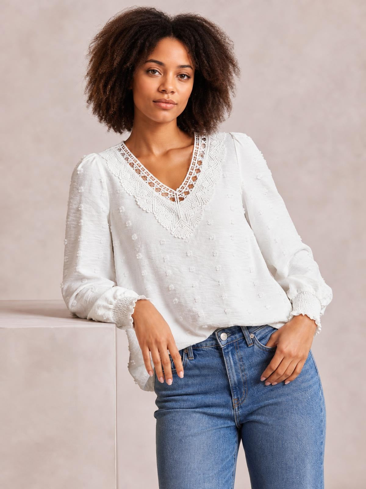 A model wears a white Swiss dot blouse with a lace-trimmed V-neck, long sleeves, gathered cuffs, and a relaxed silhouette while leaning casually.