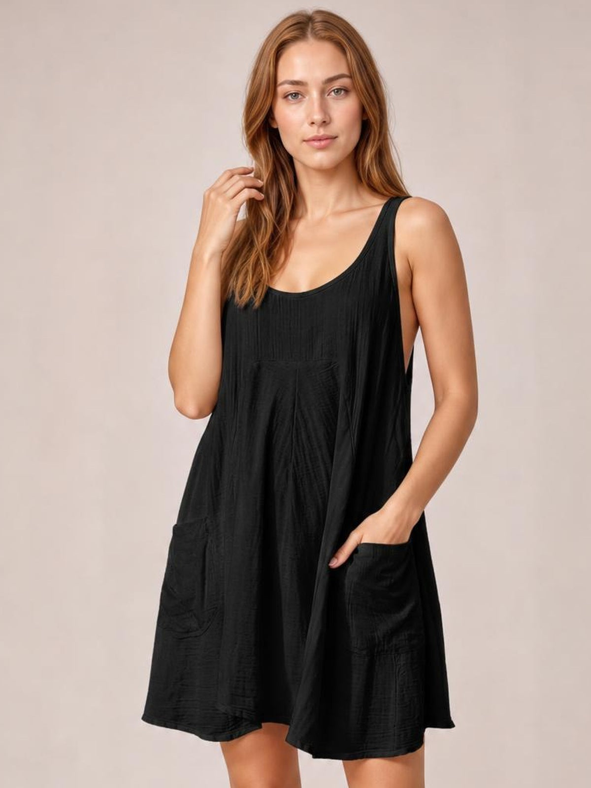 Model wearing a black scoop-neck mini dress with wide straps, a loose flowy silhouette, subtle textured fabric, and large front pockets, posing with one hand in a pocket.