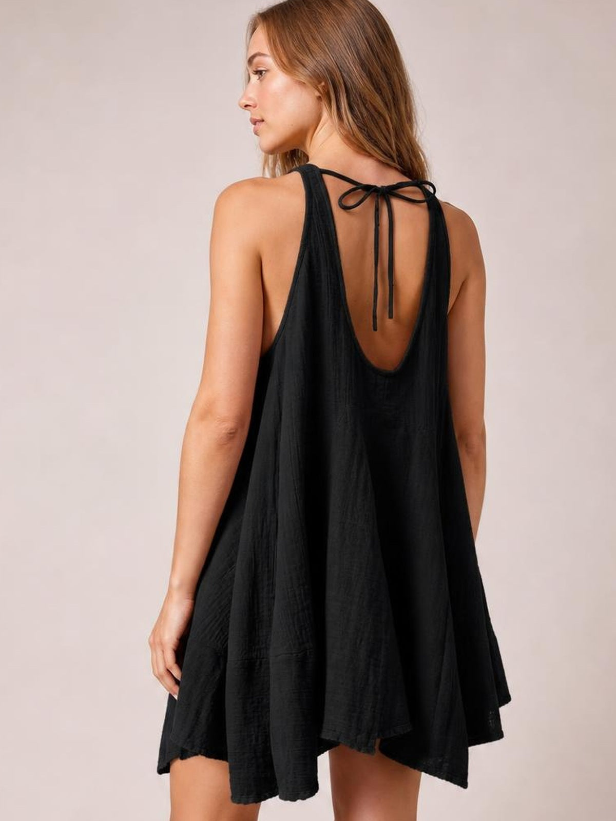 A model poses in a black tied scoop neck mini dress with wide straps, a low back, and a loose, flowing A-line silhouette.