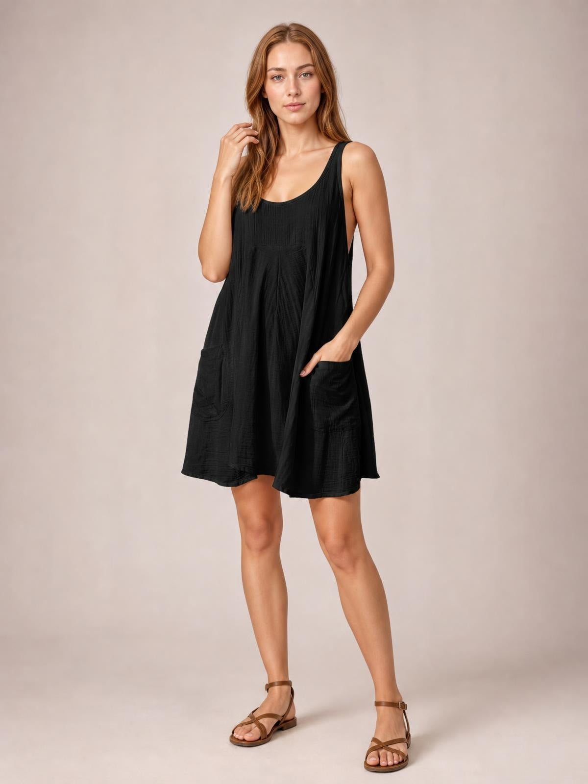 A model poses in a black tied scoop neck mini dress with wide straps, a loose A-line silhouette, subtle texture, and two front patch pockets.