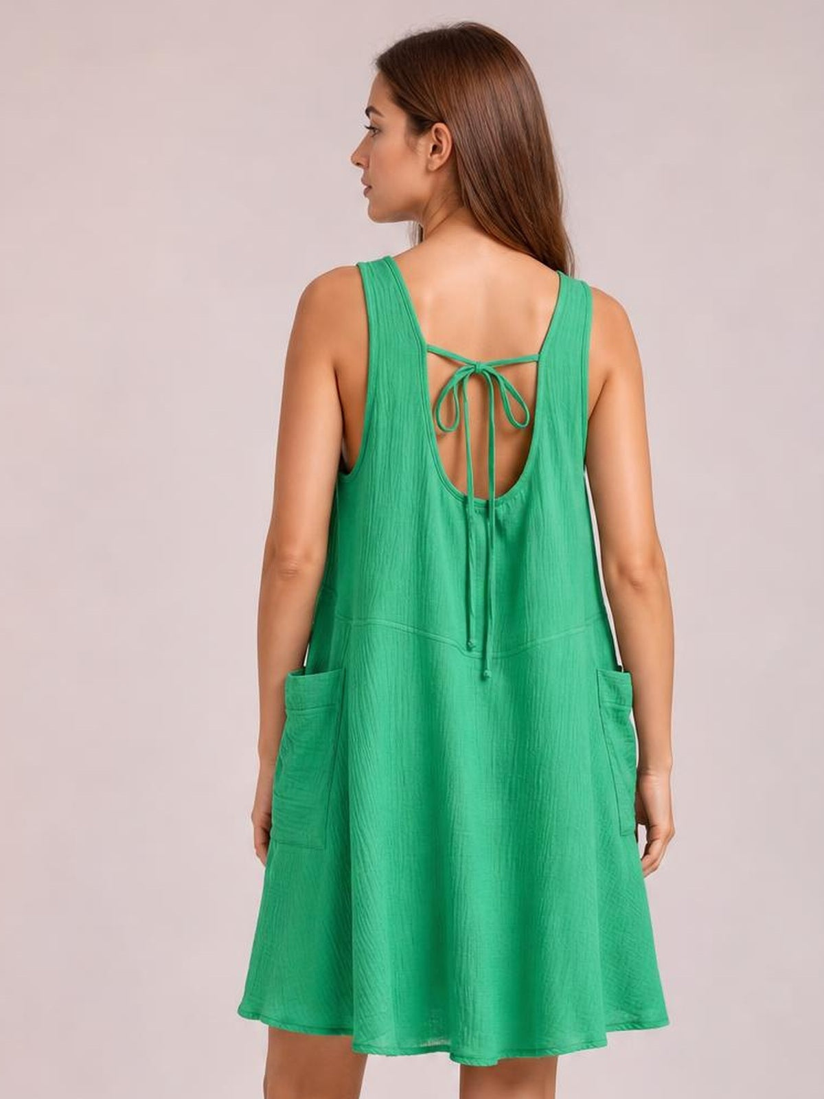 Model shown from the back in a green wide-strap mini dress with a tied open scoop back, relaxed A-line silhouette, and side pockets.