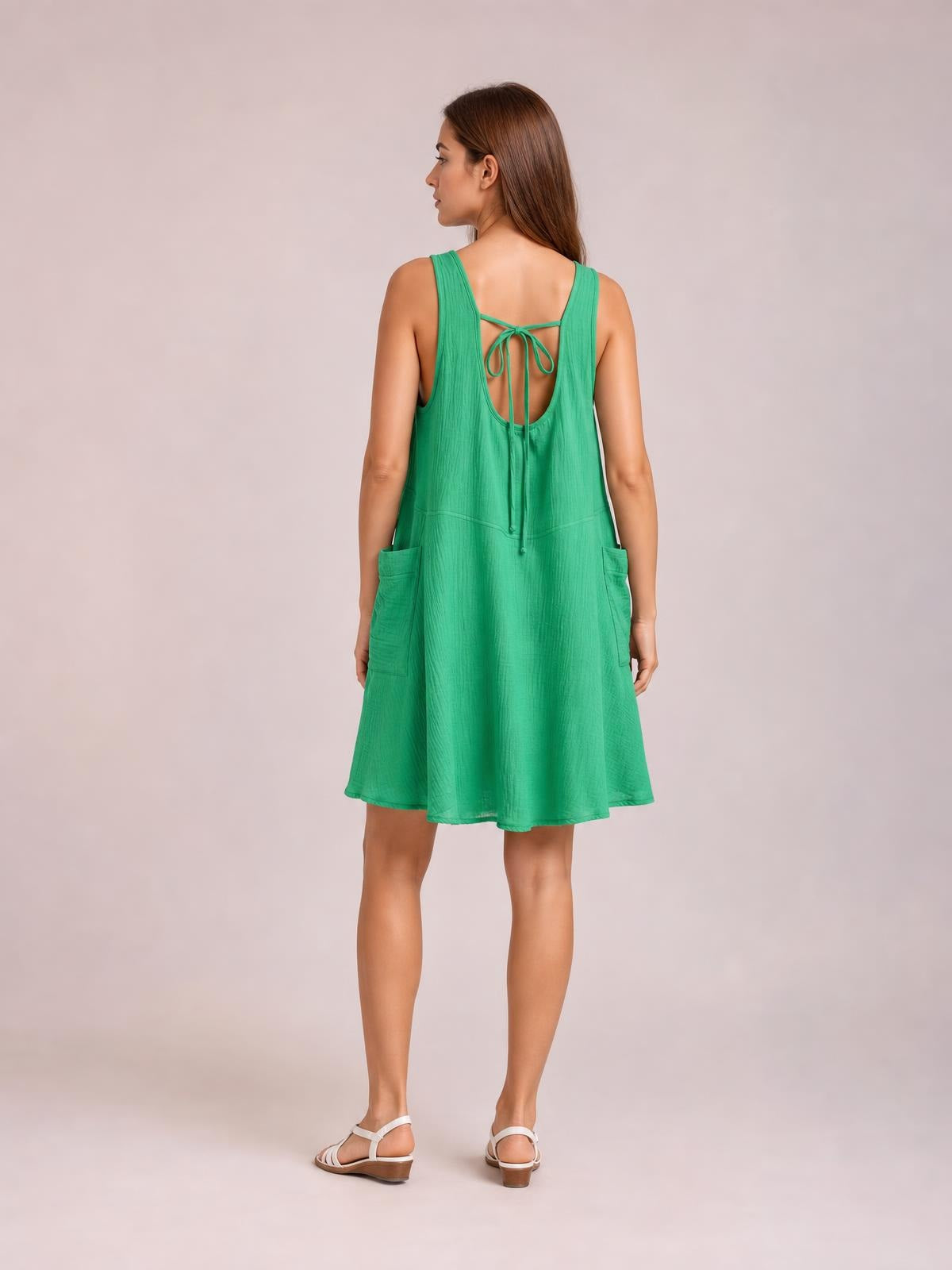 Model viewed from behind wearing a green wide-strap mini dress with a tied open back, relaxed A-line silhouette, seam detail, and side pockets.