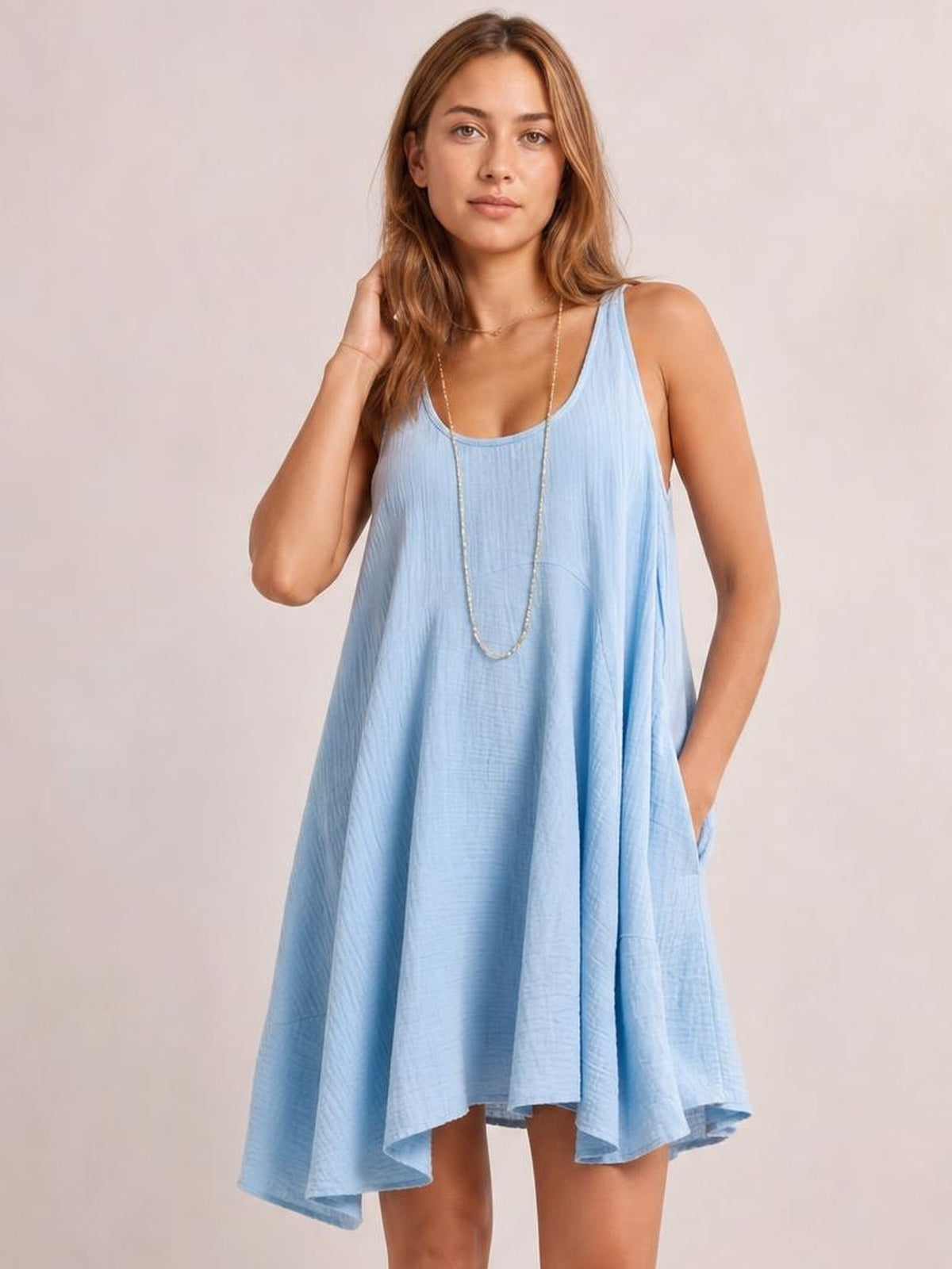 Model wearing a light blue scoop-neck mini dress with wide straps, a loose flowing silhouette, and side pockets, posing with one hand in her hair.