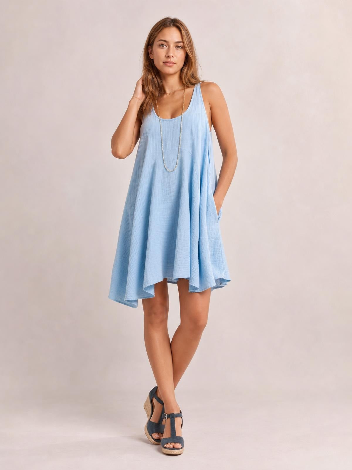 A model poses in a light blue scoop neck mini dress with wide straps, a loose flowing silhouette, subtle texture, and side pockets.