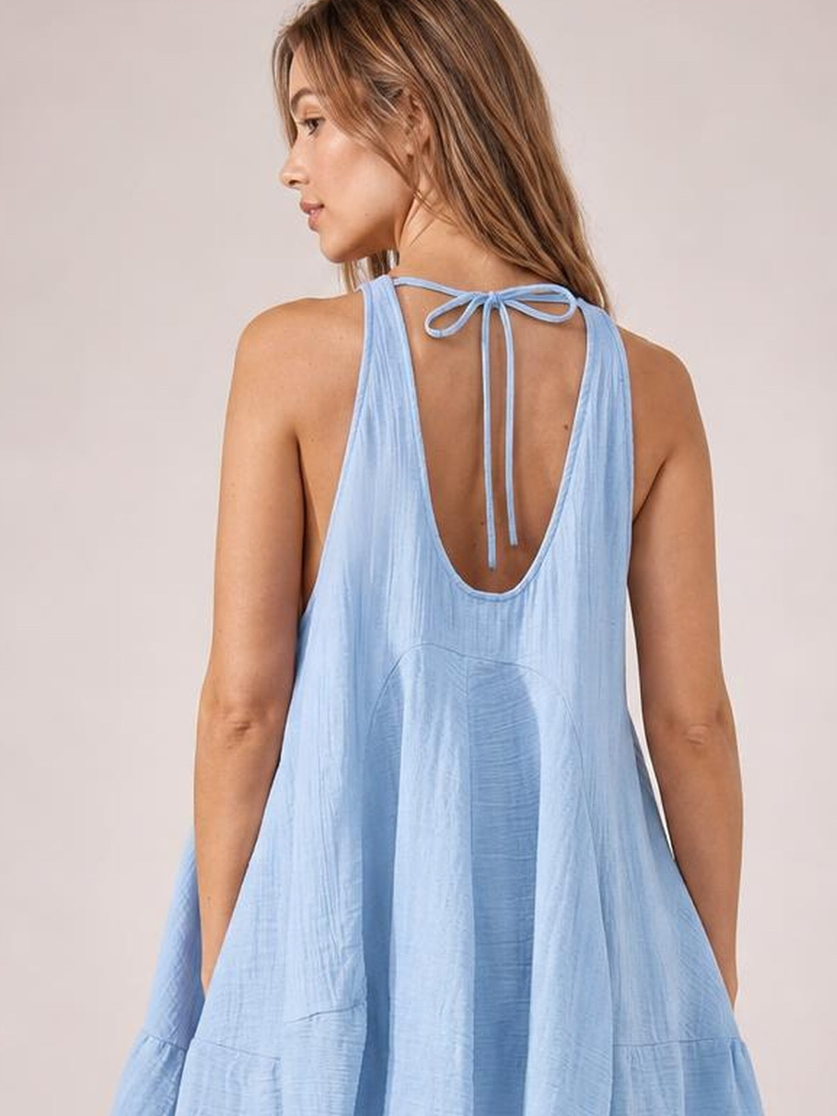 Model standing with arms relaxed, wearing a light blue tied scoop-neck mini dress with wide straps, an open low back, and a loose flowing silhouette.