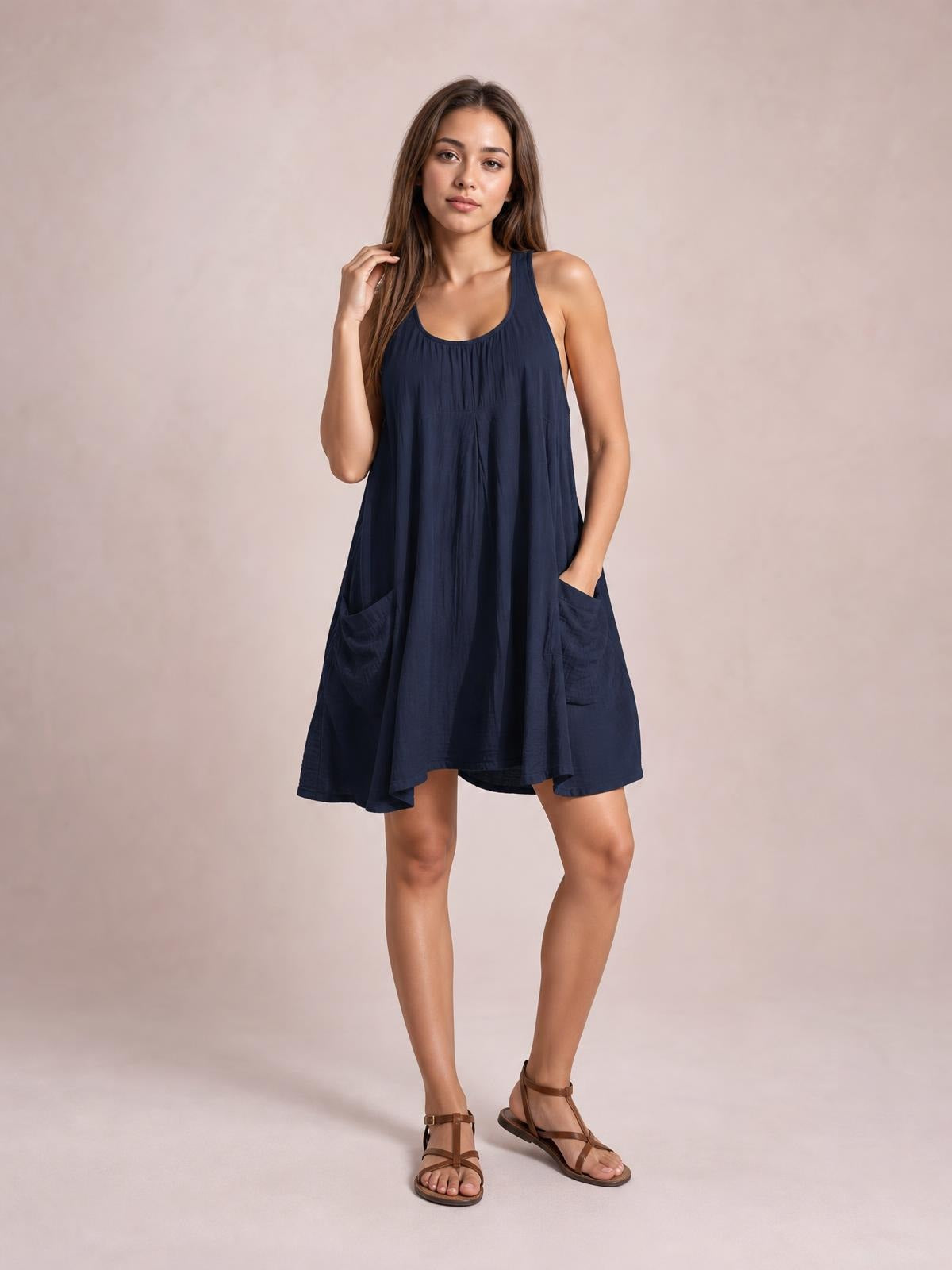 A model poses in a navy scoop-neck mini dress with wide straps, a relaxed flowing silhouette, gathered neckline, and roomy front pockets.