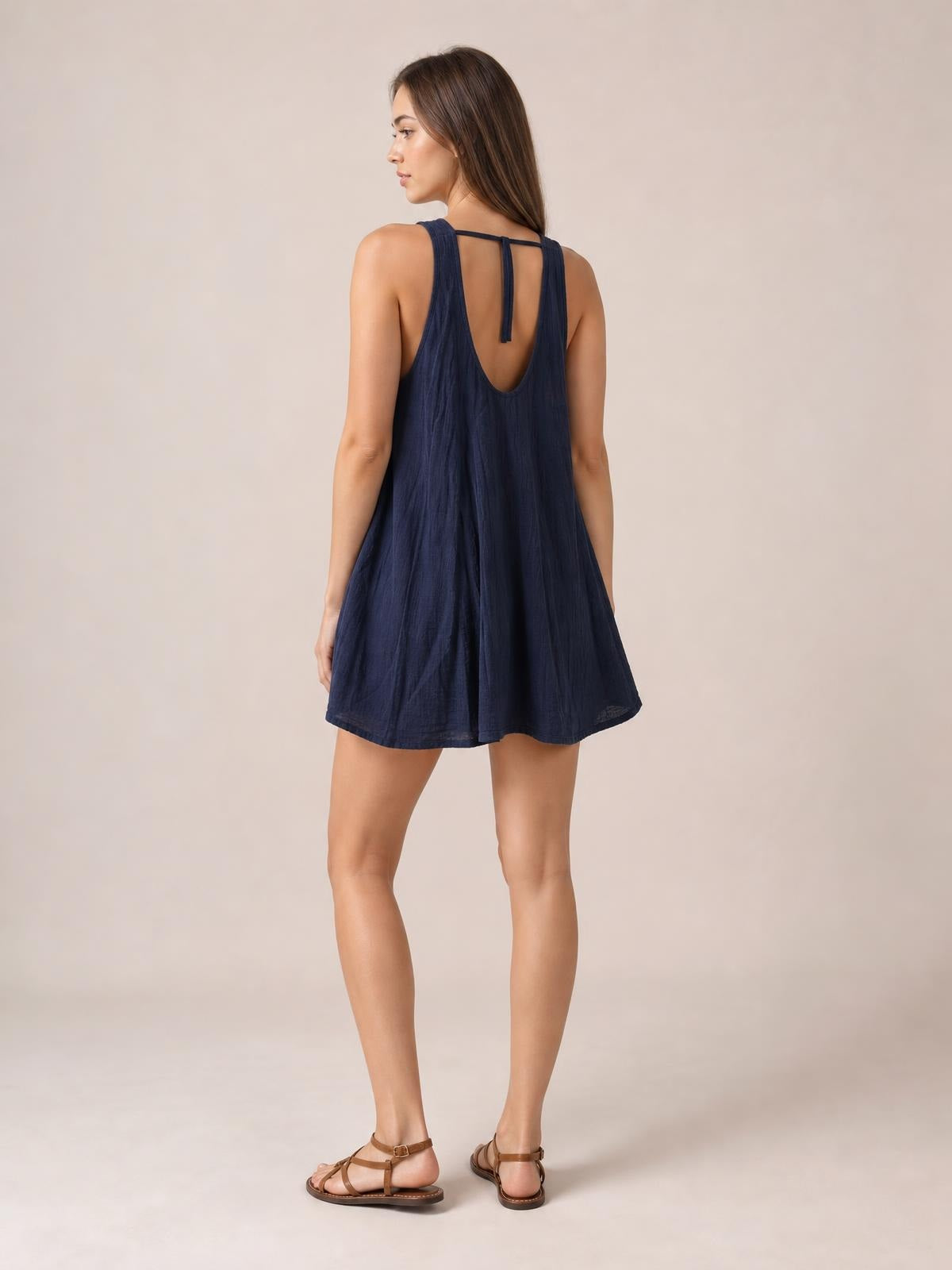 A model stands turned away in a navy tied scoop-neck mini dress with wide straps, a low scoop back, and a loose, flowing A-line silhouette.