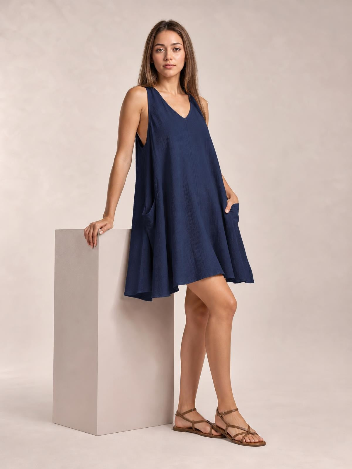A model poses in a navy scoop-neck mini dress with wide straps, a loose A-line silhouette, and side pockets, one hand resting in a pocket.