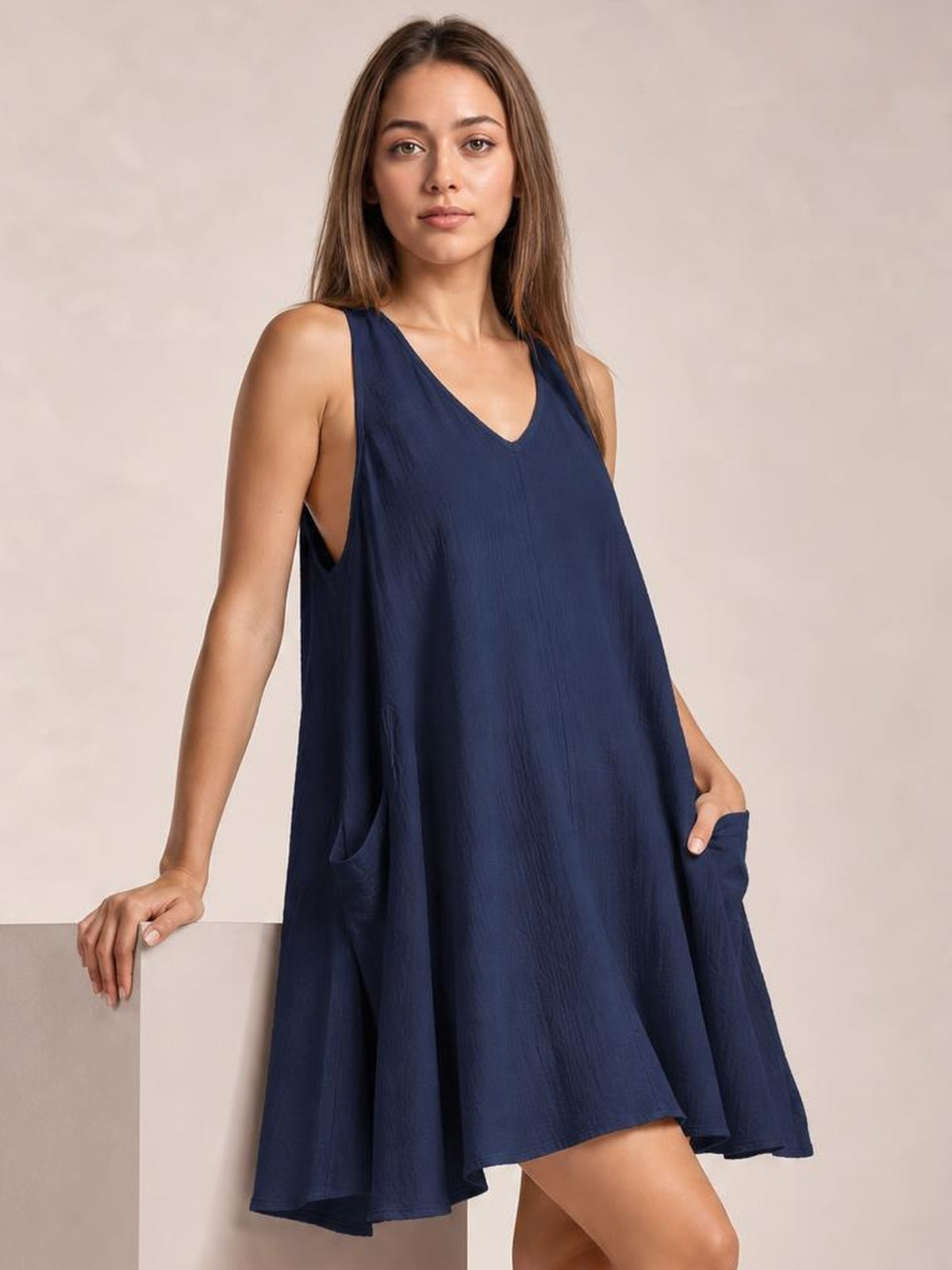 A model poses in a navy scoop-neck mini dress with wide straps, a relaxed flowing silhouette, and side pockets, resting one hand on a block.