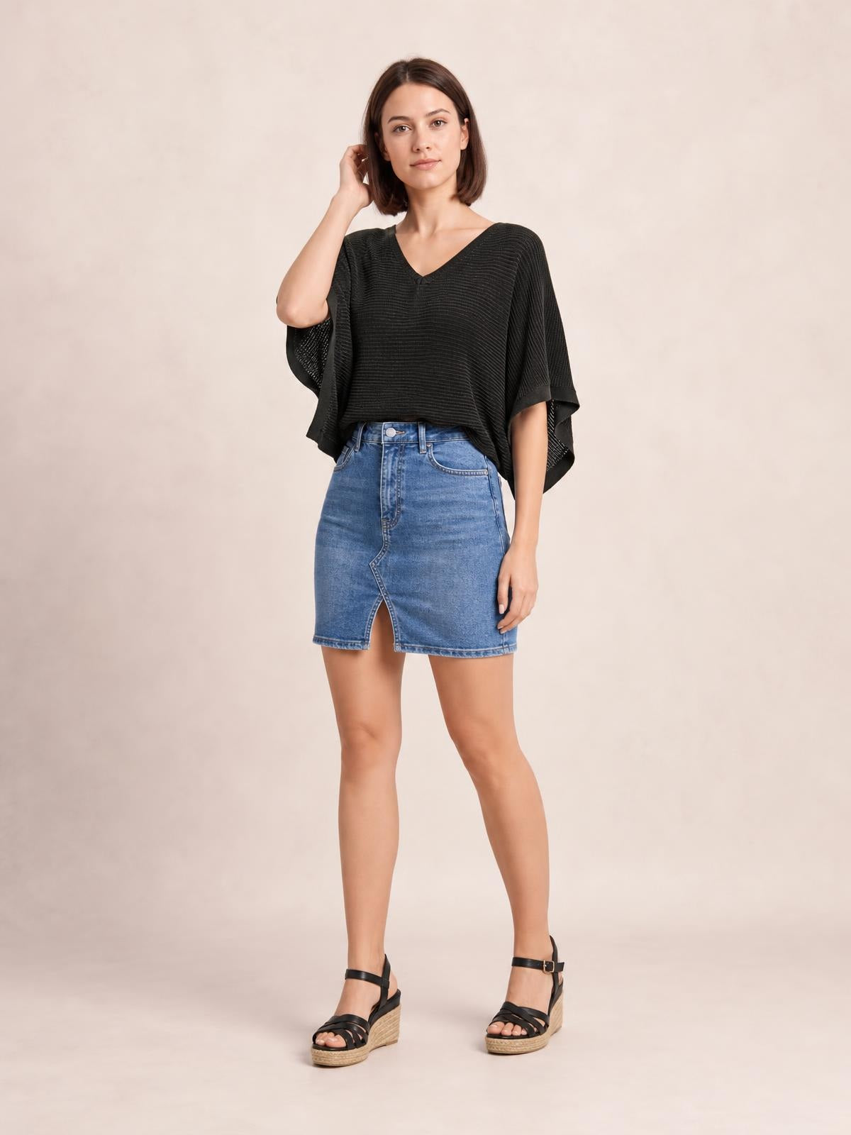 Model poses with one hand near her hair wearing a black V-neck batwing sleeve knit top with a loose draped silhouette and textured construction.