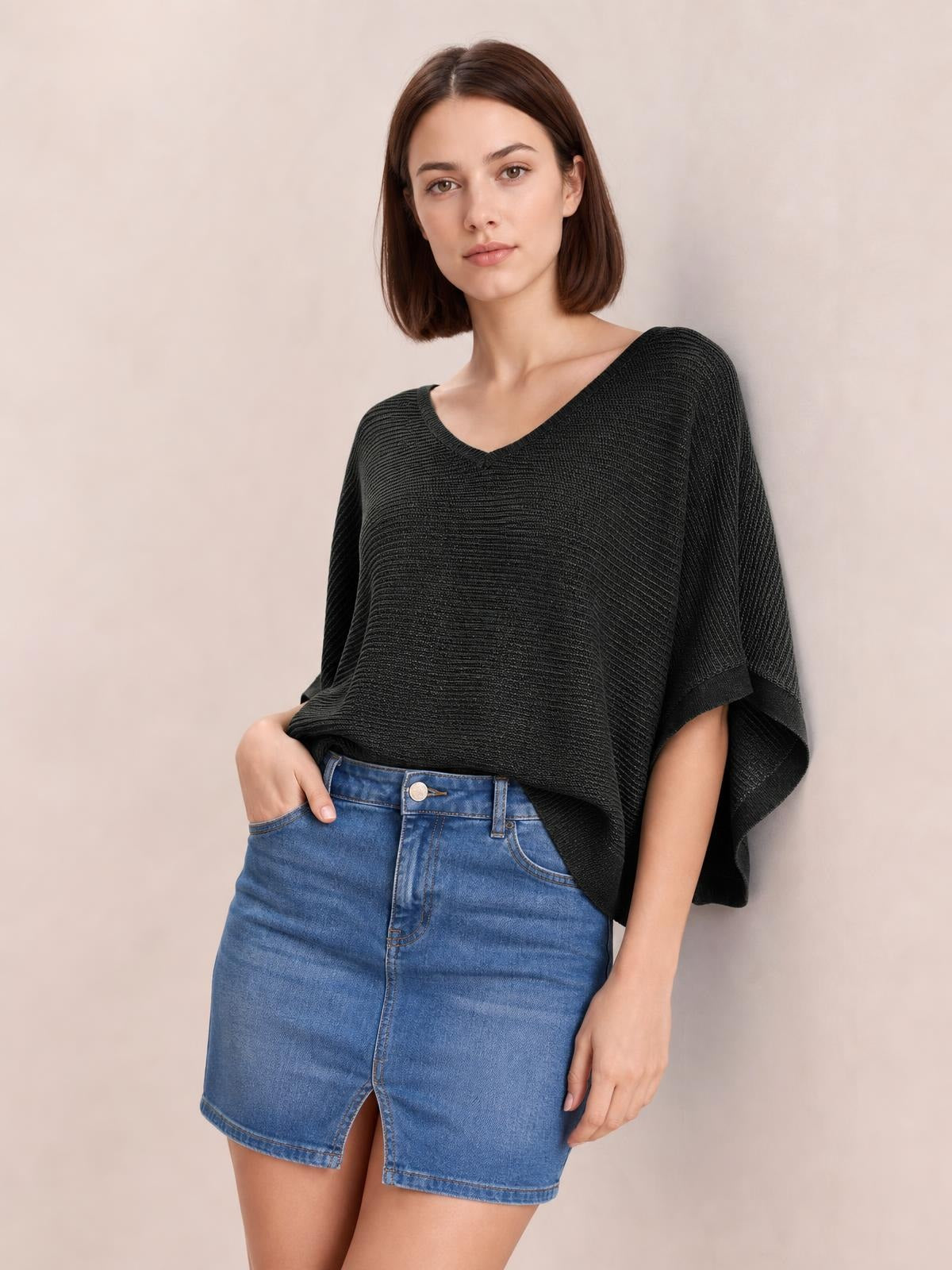 Model wearing a black V-neck batwing sleeve knit top with a loose draped silhouette and wide cuffed sleeves, paired with a denim skirt.