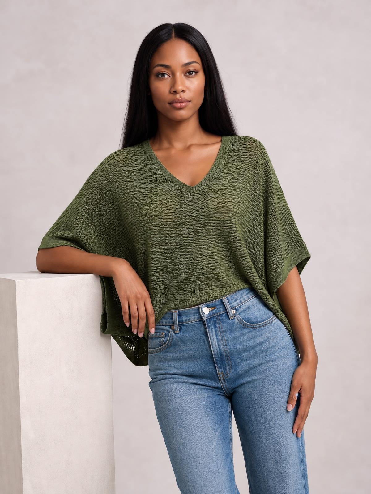 A model wears an olive green V-neck batwing sleeve knit top with a loose, draped silhouette and short cuffs, leaning casually with one arm resting beside her.