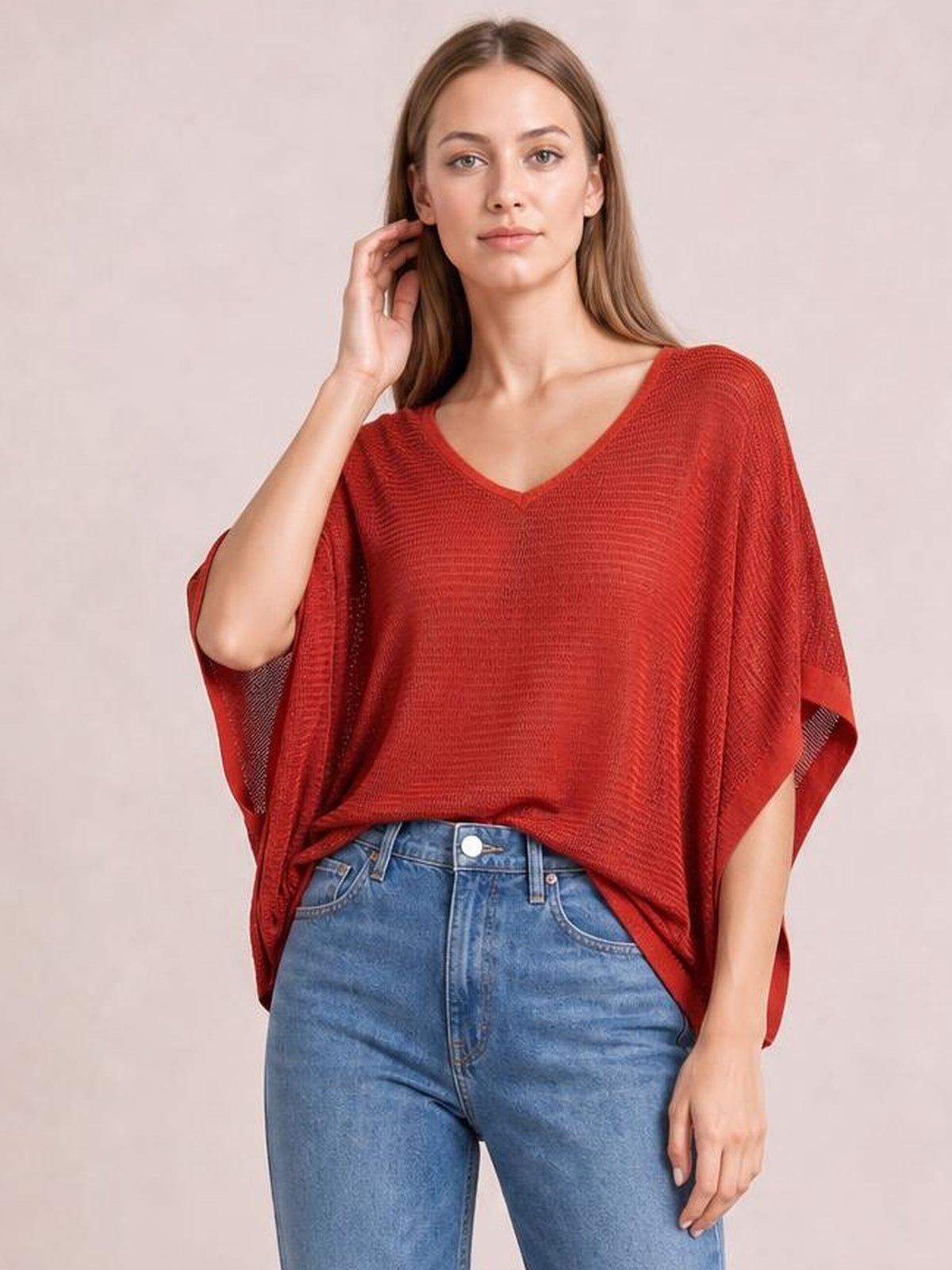 Model wearing a rust-red V-neck batwing sleeve knit top with a loose draped silhouette and wide dolman sleeves, paired with blue jeans.