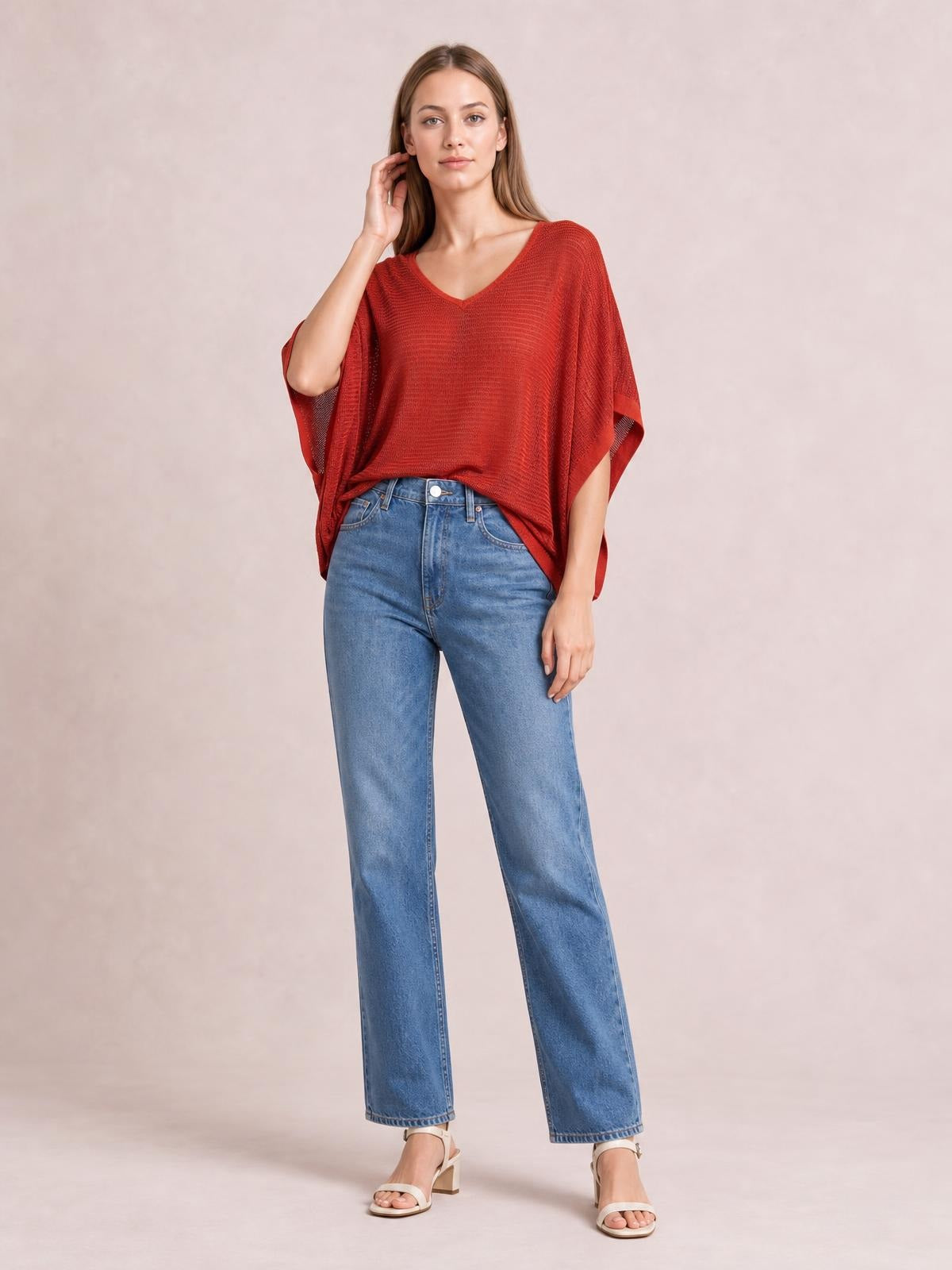 Model wearing a red V-neck batwing sleeve knit top with a loose draped silhouette and side slits, styled with straight-leg jeans and heels.