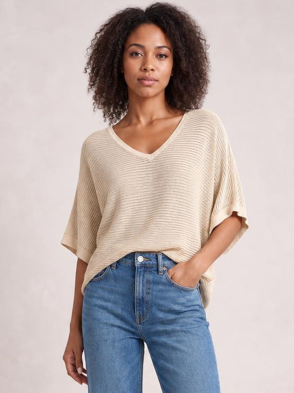 Model wears a cream V-neck batwing sleeve knit top with a loose, draped silhouette, open-knit texture, and wide short sleeves, paired with jeans.