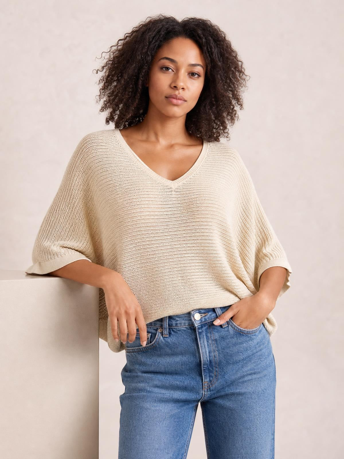 A model leans casually with one hand in her jeans pocket, wearing a cream V-neck batwing sleeve knit top with a relaxed, loose silhouette.