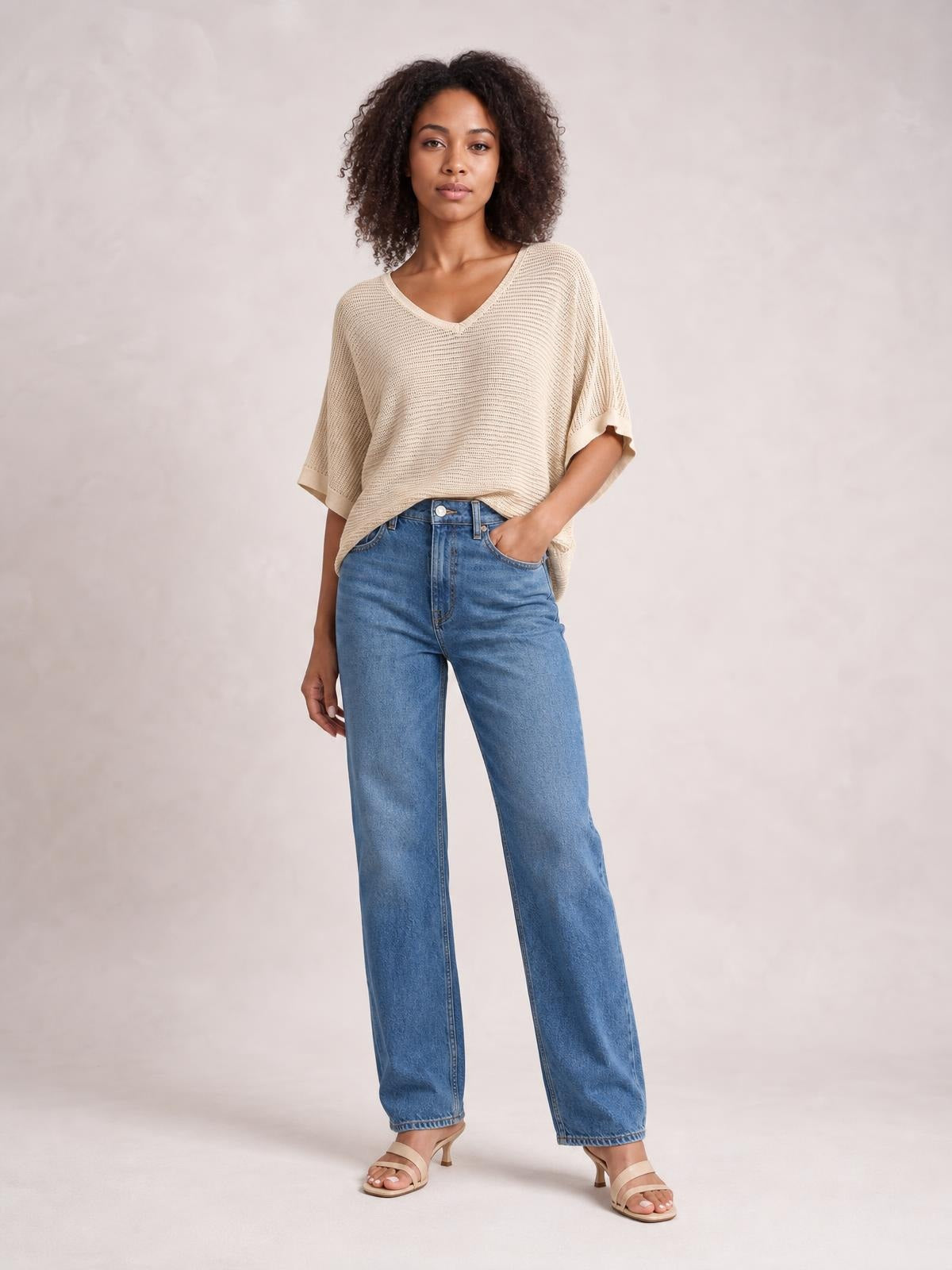 Model wearing a beige V-neck batwing sleeve knit top with a relaxed silhouette, dropped shoulders, and rolled cuffs, standing with one hand in her jeans pocket.