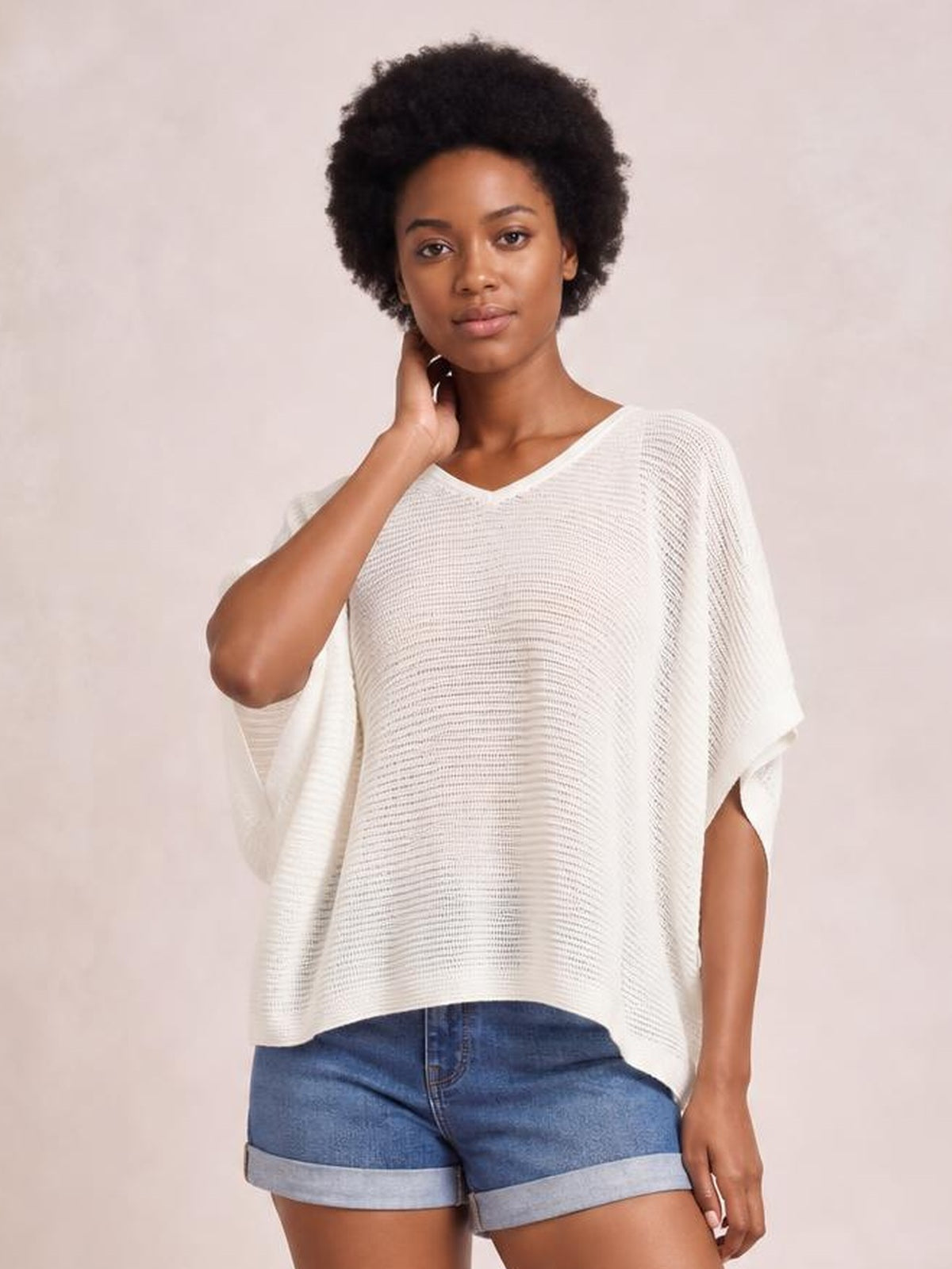 Model poses with one hand at her neck wearing a white V-neck batwing sleeve knit top with a relaxed, draped silhouette and ribbed texture.