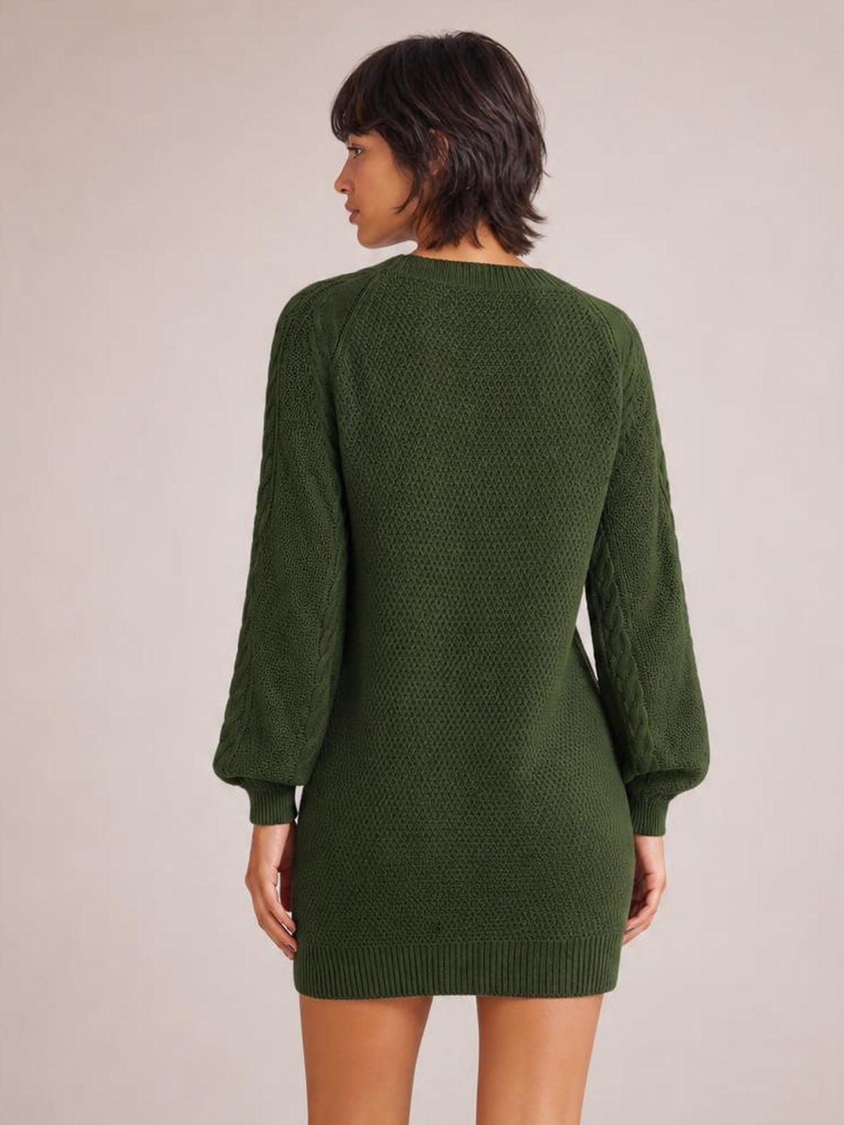 Model shown from behind in a forest green diamond cable-knit sweater dress with long sleeves, ribbed cuffs and hem, and a relaxed straight silhouette.