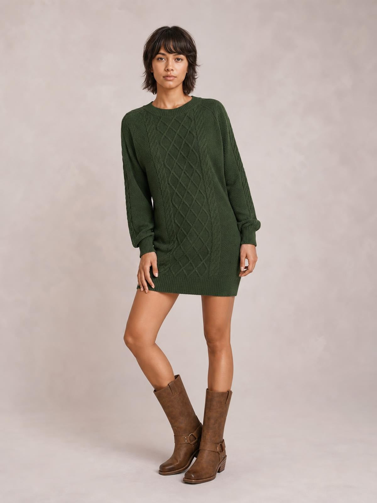 Model poses in a dark green diamond cable-knit sweater dress with long sleeves, a crew neckline, ribbed cuffs and hem, and a relaxed mini silhouette.