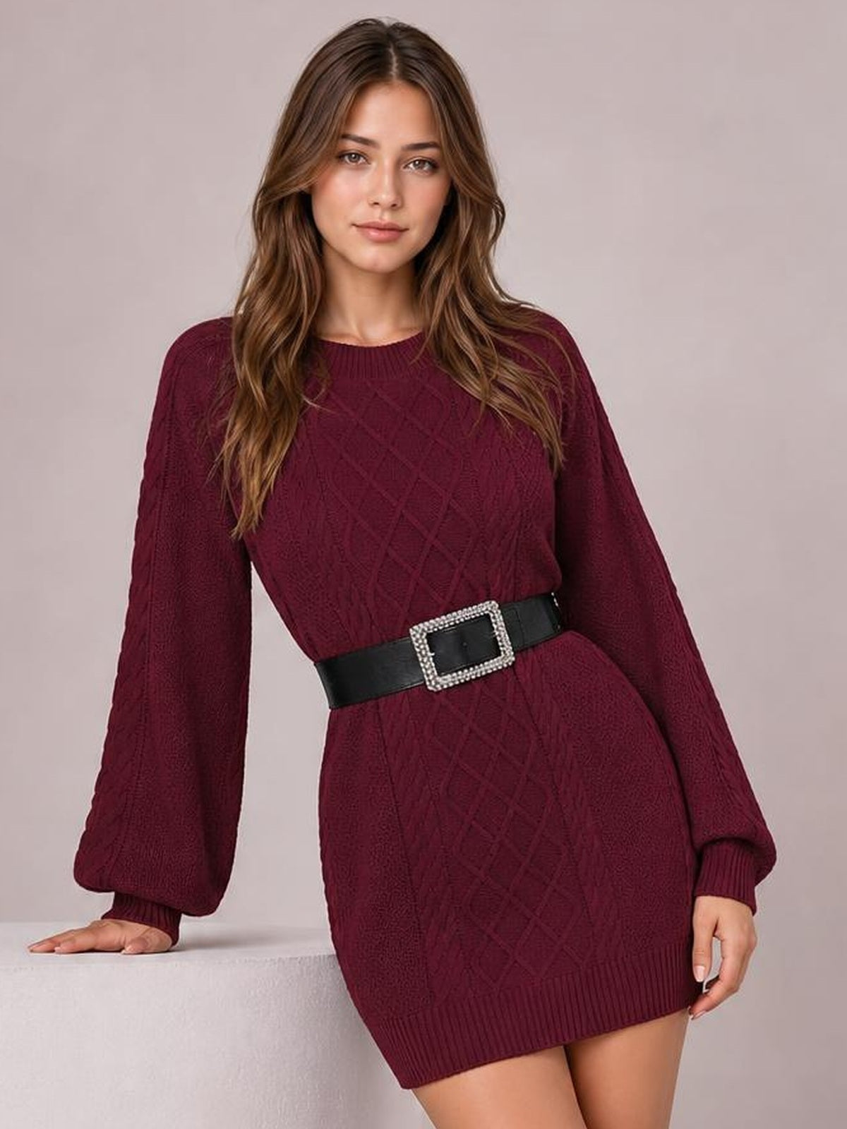 A model leans with one hand resting beside her, showcasing a burgundy diamond cable-knit sweater dress with long sleeves, ribbed trim, and a short fitted silhouette.