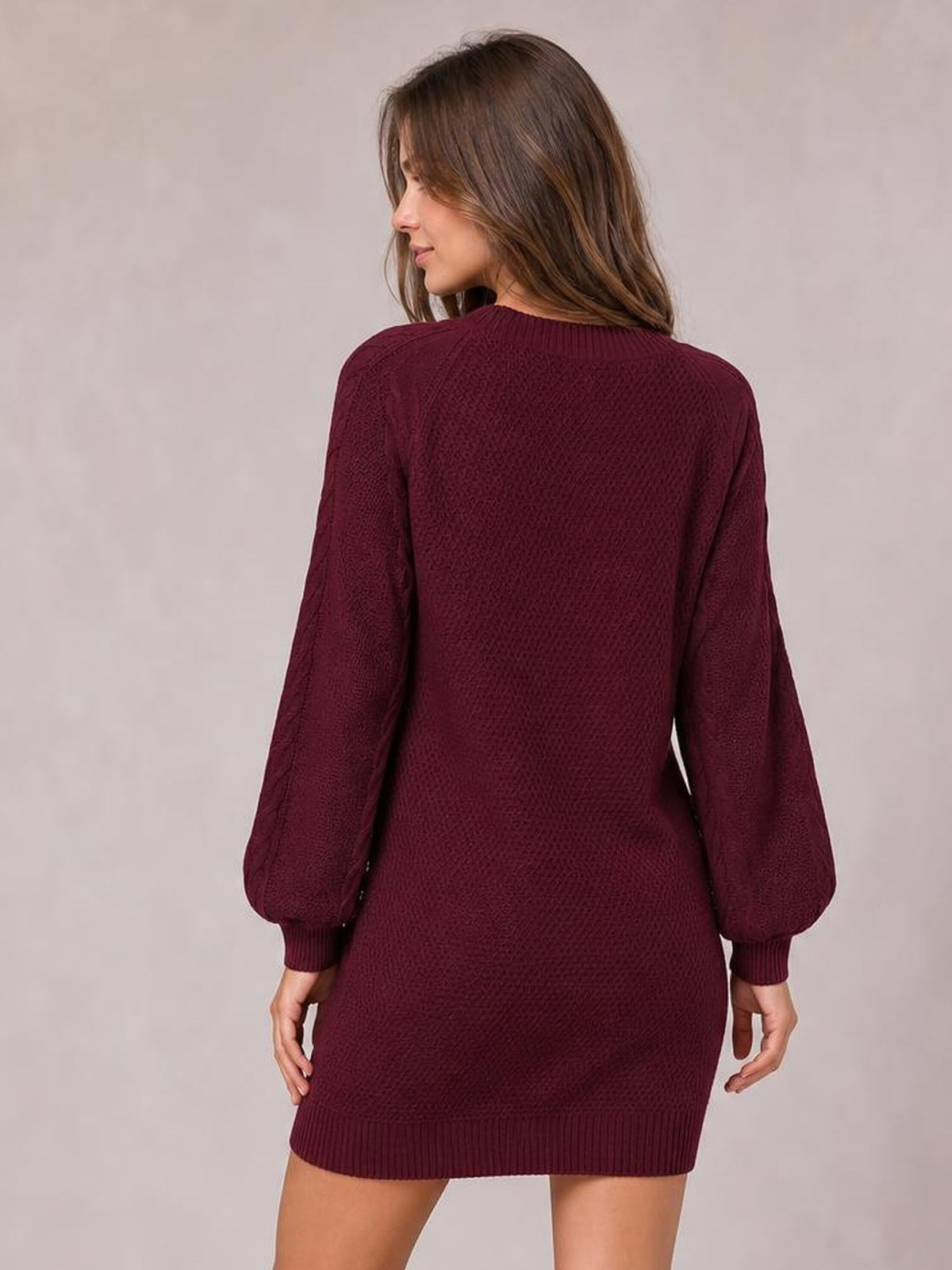 A model turns slightly to show the back of a burgundy diamond cable-knit sweater dress with long sleeves, ribbed cuffs, neckline, and hem.