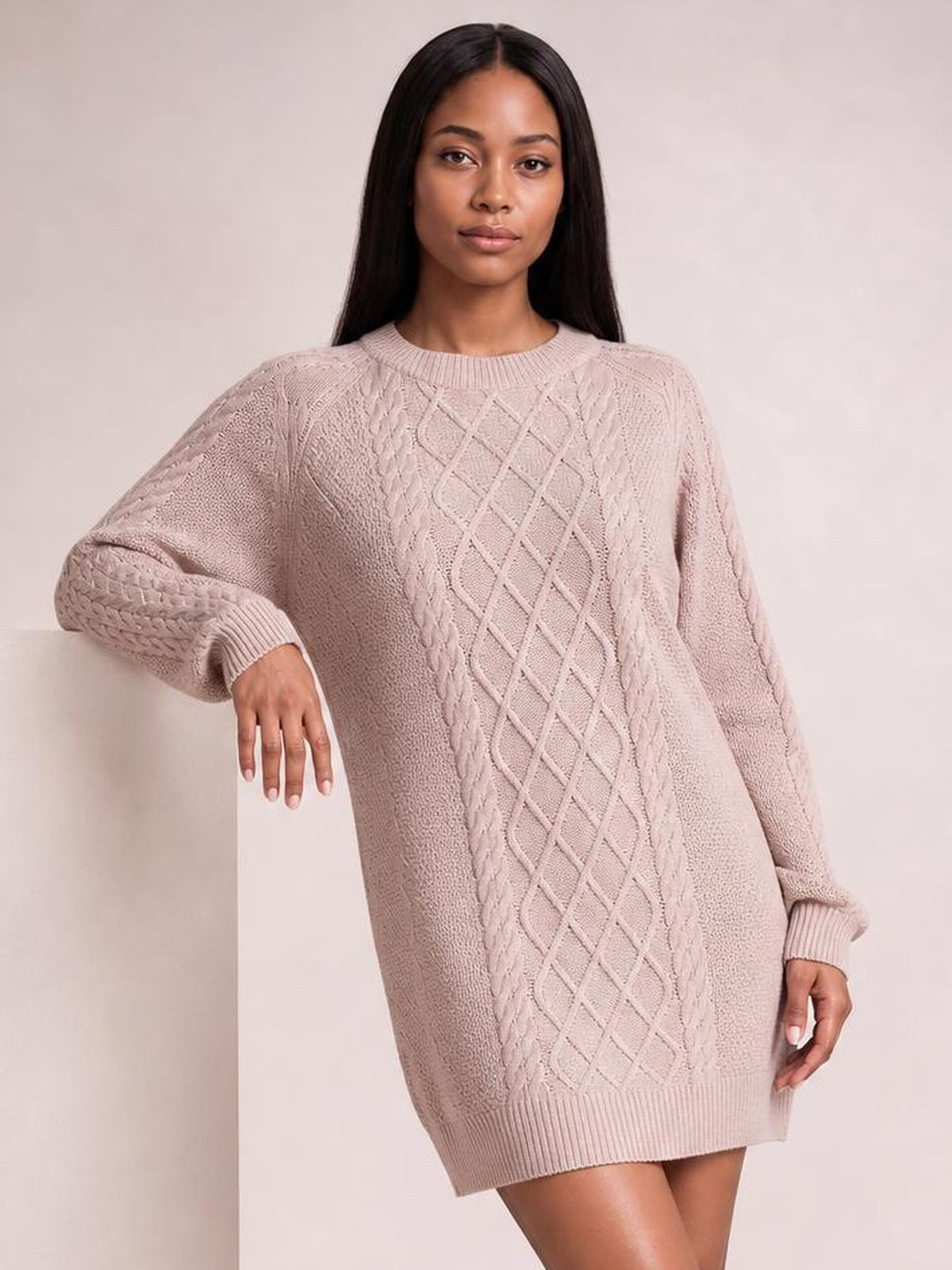 Model in a soft blush diamond cable-knit sweater dress with long sleeves, ribbed crew neckline, cuffs, and hem, shown in a relaxed leaning pose.