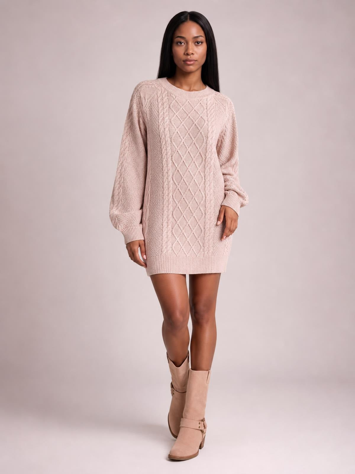 Model stands facing forward in a blush pink diamond cable-knit sweater dress with long sleeves, ribbed cuffs and hem, and a relaxed straight silhouette.
