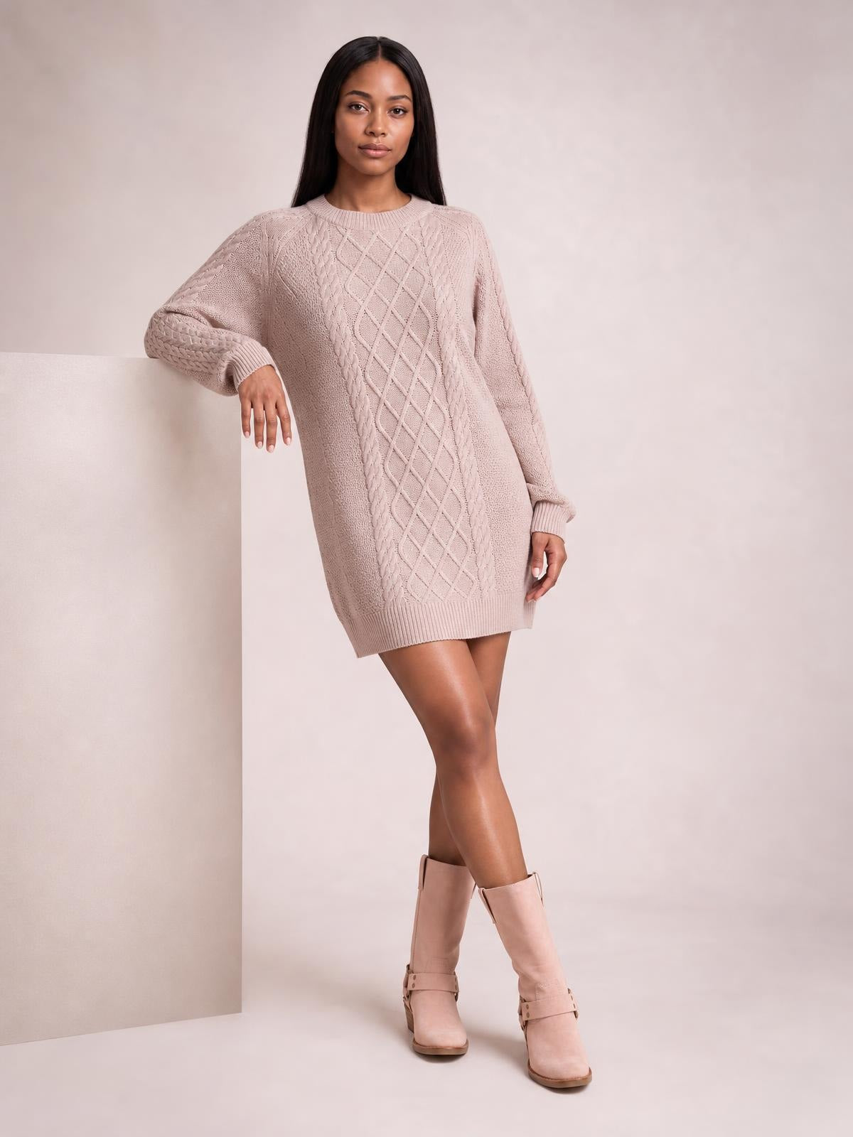 Model poses in a blush pink diamond cable-knit sweater dress with long sleeves, ribbed crew neckline and hem, relaxed silhouette, and crossed legs.