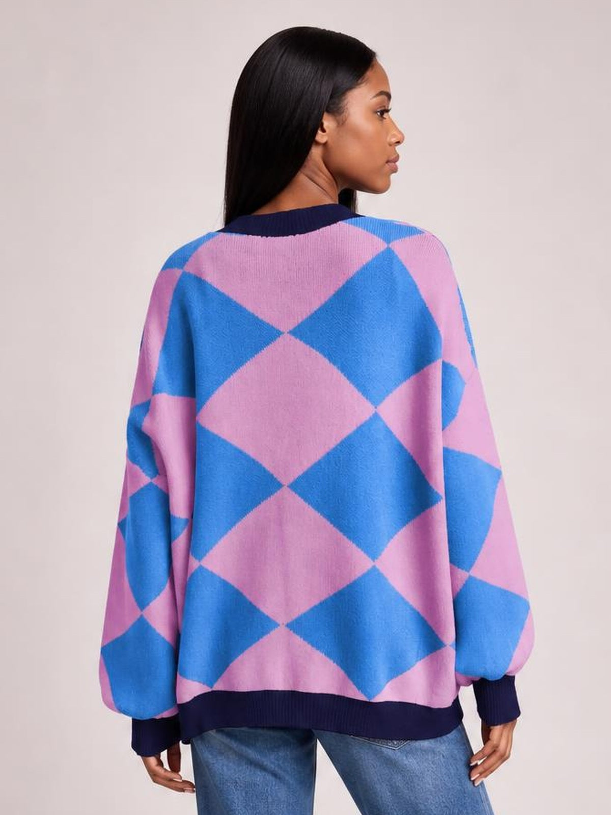 A model turns to show the relaxed silhouette of a pink and blue diamond-pattern button-up knit cardigan with ribbed navy trim and long sleeves.